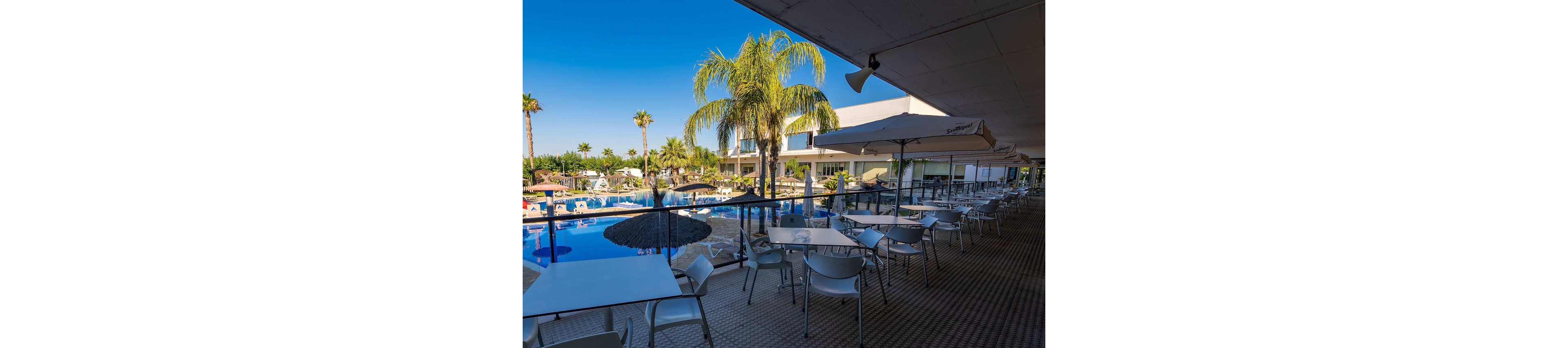 An outdoor hotel terrace with white tables and chairs overlooks a large swimming pool. Sun loungers and umbrellas surround the pool area, framed by tall palm trees. Modern resort buildings stand in the background under a clear blue sky. The scene suggests a relaxed vacation atmosphere in a warm, sunny destination.