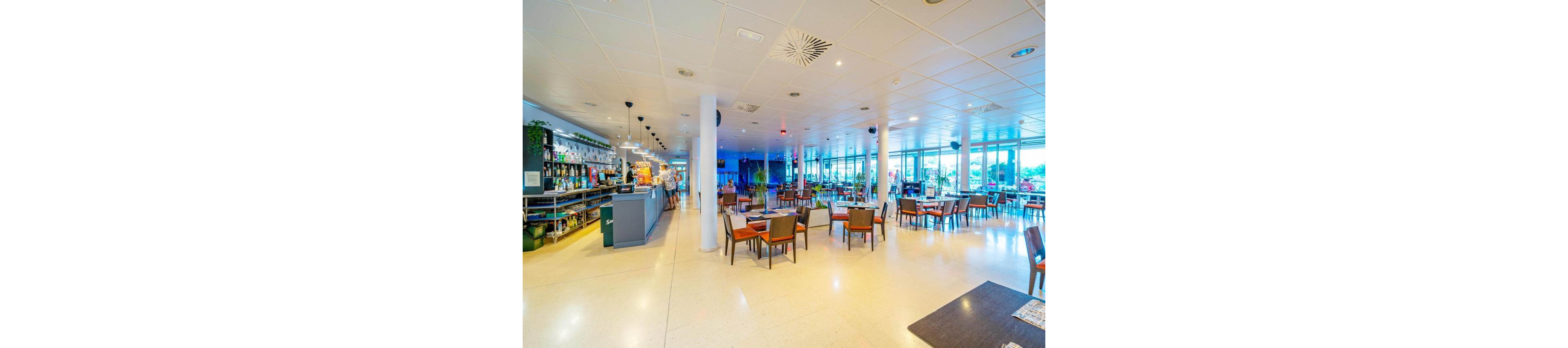 Spacious contemporary restaurant interior with a bar counter on the left and neatly arranged wooden tables and chairs throughout the room. Large floor-to-ceiling windows on the right let in abundant natural light and offer views of the outside. The ceiling features recessed lighting and ventilation panels, creating a clean, modern look. A few people are visible near the bar area, while most of the seating area remains empty and ready for guests.