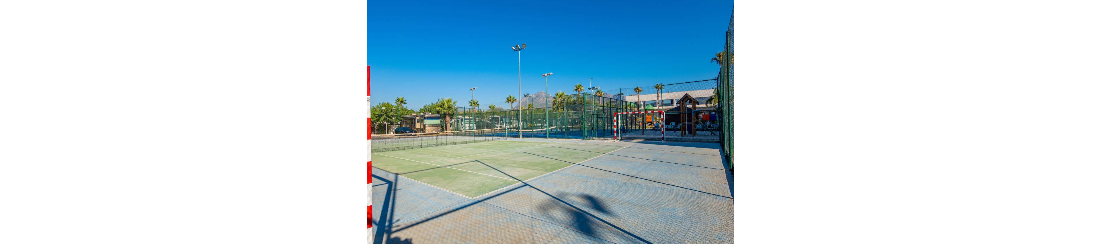 An outdoor sports complex features adjacent courts enclosed by tall fencing. A goalpost stands on a tiled surface next to a green playing court, suggesting multiple sports activities. Palm trees line the perimeter, and buildings and mountains are visible in the background. Bright daylight and a cloudless blue sky create a vibrant, summery atmosphere.