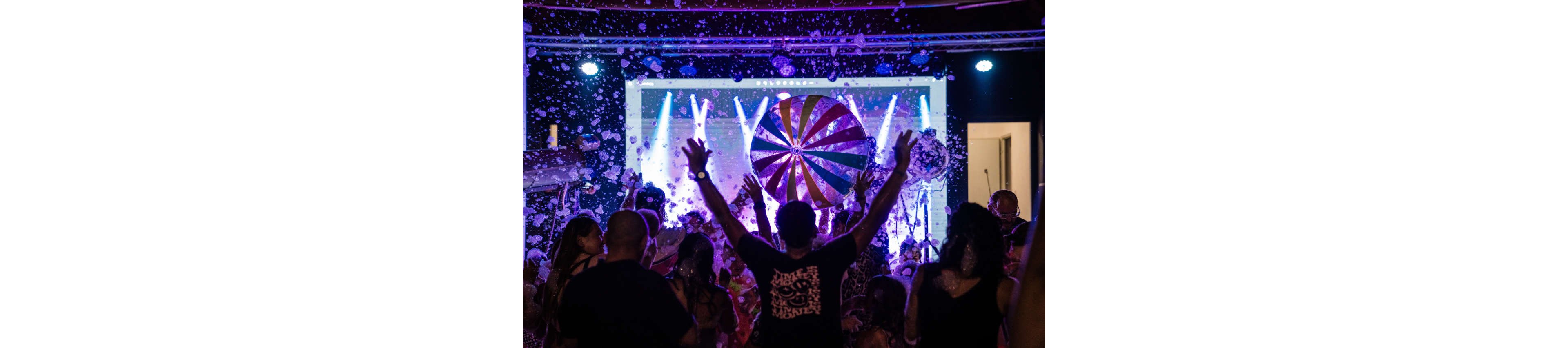 A lively crowd raises their hands toward a large colorful beach ball at a foam party. The scene is set indoors in front of a bright stage with purple and blue lights. Soap bubbles and foam fill the air, creating a festive and energetic atmosphere. People of various ages enjoy the music and playful environment together.