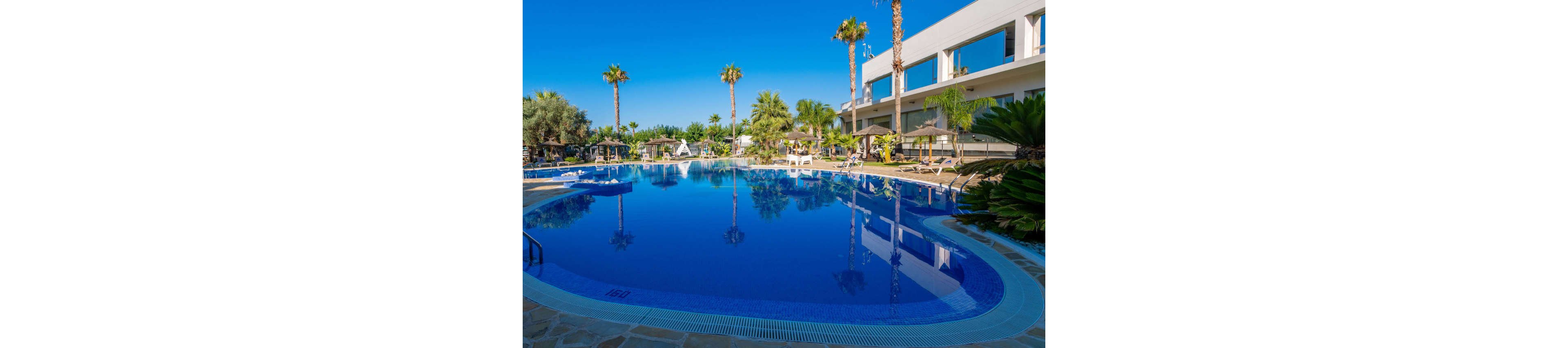 A large, curved swimming pool reflects a clear blue sky at a modern resort. White sun loungers and umbrellas line the poolside, surrounded by lush palm trees and tropical plants. A contemporary white building with balconies overlooks the water, adding to the upscale atmosphere. The scene is bright and tranquil, suggesting a relaxing vacation setting.