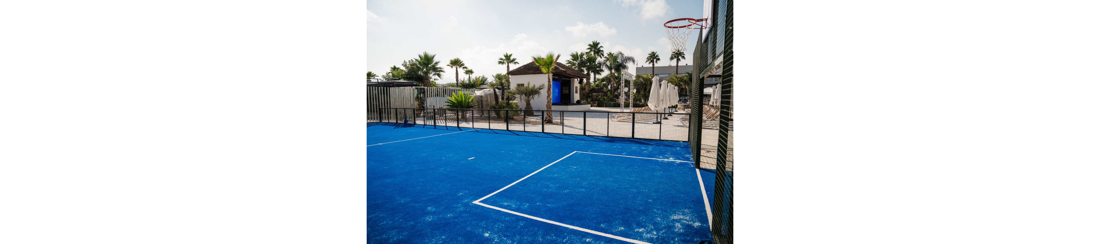An outdoor blue sports court is shown beside a fenced recreation area. A basketball hoop is mounted on the right side of the court. In the background, there is a poolside area with umbrellas, lounge chairs, and a small building. Palm trees and clear skies create a sunny, vacation-like atmosphere. The scene suggests a modern resort or residential complex with leisure facilities.