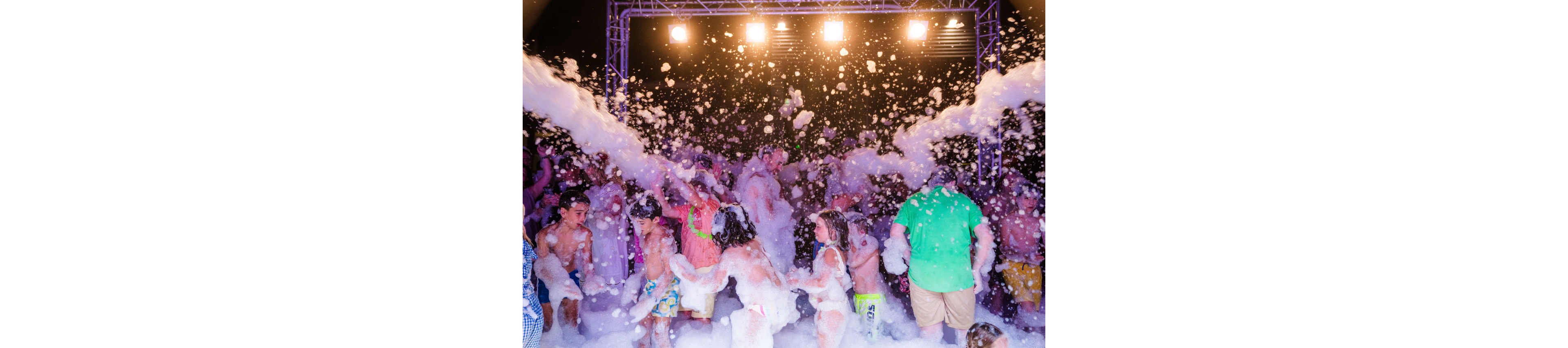 A large group of children and a few adults play in thick foam at a lively party. The scene is set under bright stage lights and a metal truss structure at night. People are covered in bubbles, dancing and throwing foam into the air. The atmosphere feels energetic, festive, and carefree.
