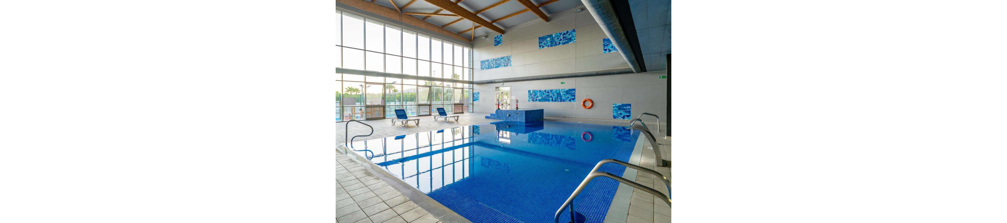 Spacious indoor swimming pool with clear blue water is shown in a modern leisure facility. Large floor-to-ceiling windows let in natural light and reveal palm trees outside. Blue lounge chairs line the tiled deck, and a raised platform stands at the far end of the pool. Safety equipment, including a life preserver and handrails, is visible along the walls and pool edges.