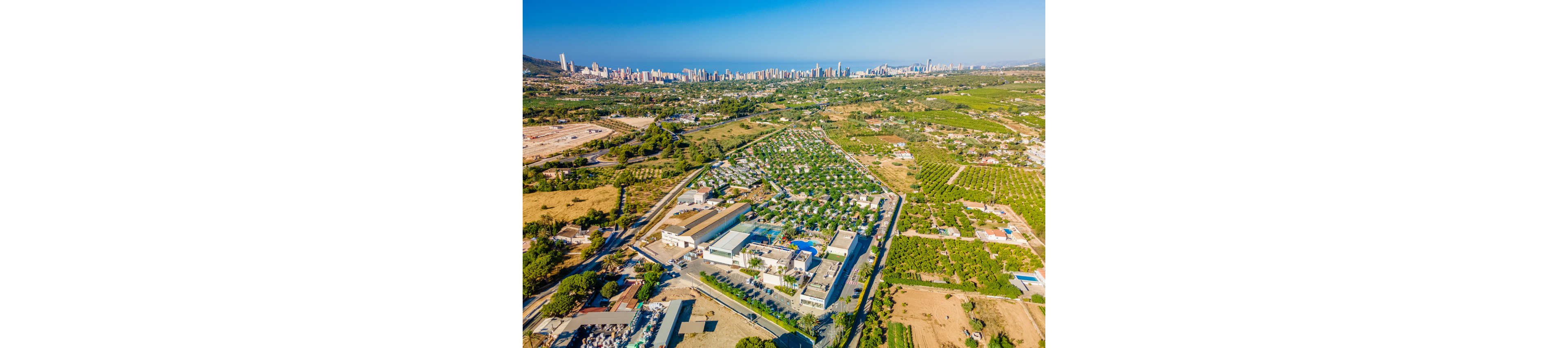 High-angle aerial view shows a large resort or campground complex surrounded by green fields and orchards. Numerous small units or cabins are arranged in neat rows with roads and pathways connecting the property. In the distance, a dense modern city skyline stretches along the coastline beside the sea. The scene is captured on a clear sunny day with bright blue sky and vivid colors. The contrast between rural farmland and urban high-rises highlights diverse land use in the coastal region.