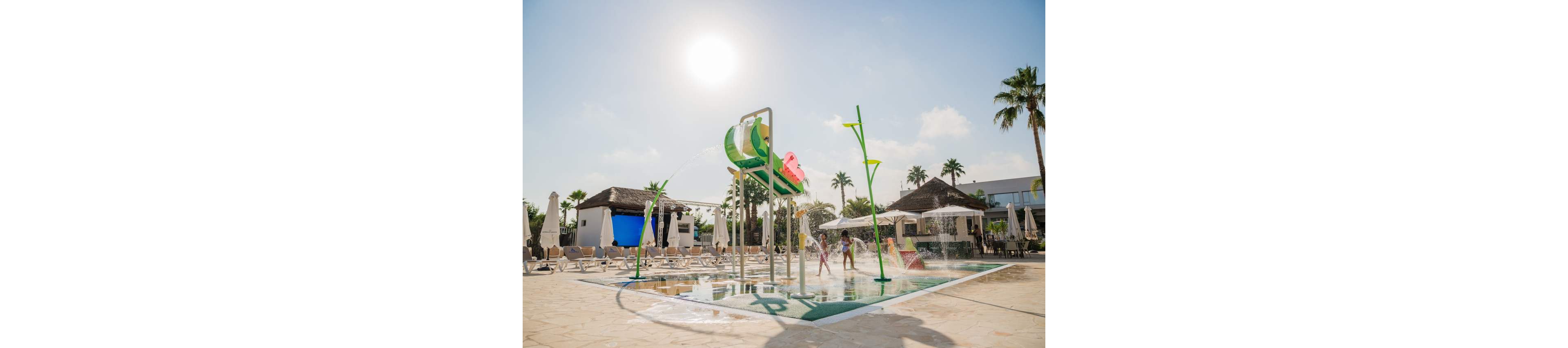 Children play under colorful water features at an outdoor splash park in a sunny resort setting. The shallow water area includes spraying arches, tipping buckets, and fountains surrounded by lounge chairs and umbrellas. Palm trees and low resort buildings frame the background under a bright midday sun. The scene conveys a warm, family-friendly vacation atmosphere focused on fun and relaxation by the pool.