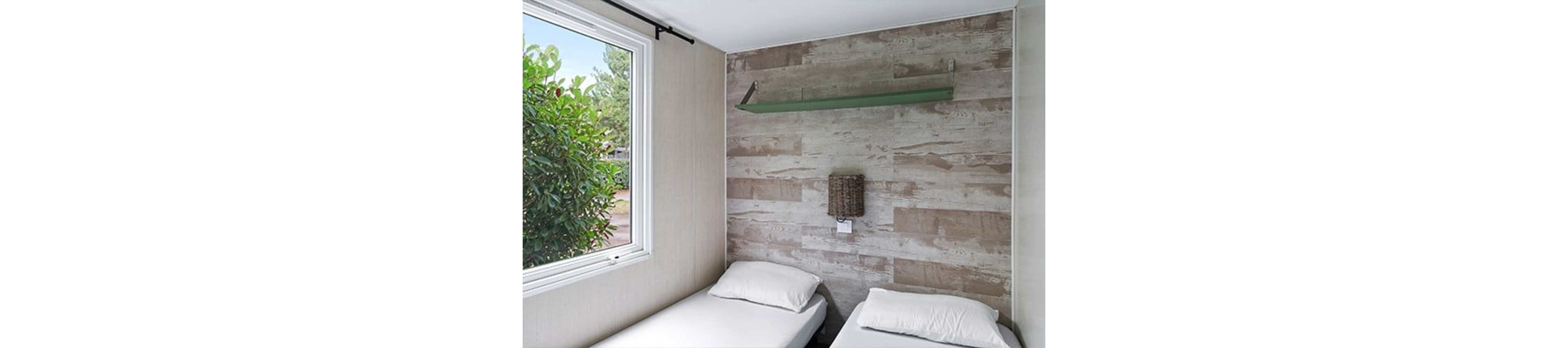 A compact bedroom features two single beds with white bedding placed against a wood-textured accent wall. A woven wall-mounted light fixture is centered between the beds, and a minimalist glass shelf is installed above it. Natural light enters through a large window on the left, revealing green foliage outside. The overall space feels simple, bright, and functional, suitable for a guest room or vacation rental.