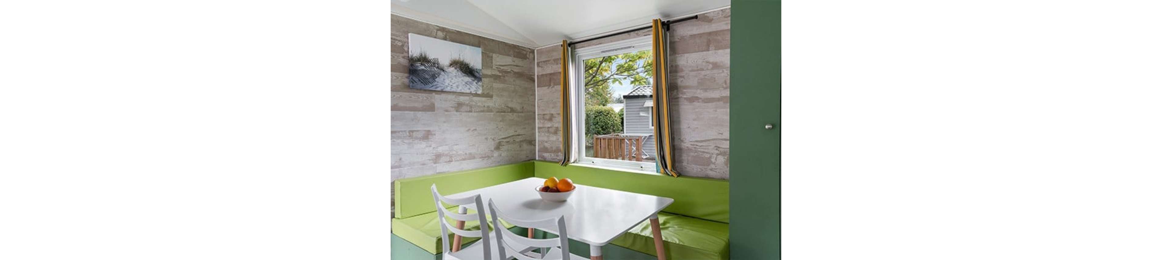 A compact dining nook features a white rectangular table surrounded by lime green bench seating and two white chairs. The space is set against light wood-paneled walls with a framed coastal photograph hanging above the bench. A large window with yellow and gray curtains lets in natural light and offers a view of a deck and neighboring building. A bowl of assorted fruit sits at the center of the table, adding a pop of color to the modern, minimalist interior.