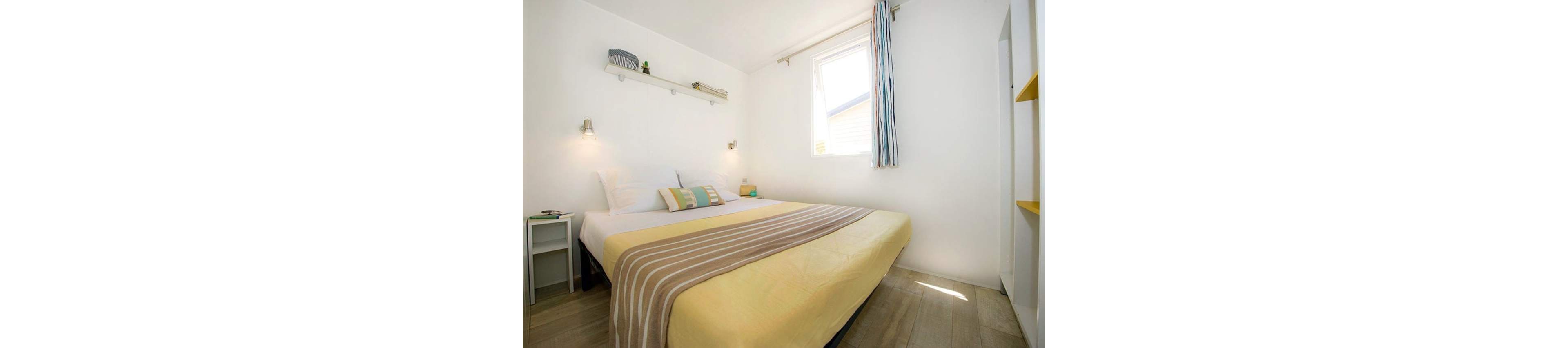 A compact, modern bedroom features a large bed with a yellow cover and striped throw blanket. Natural light streams in through a single window, illuminating the white walls and light wood flooring. Simple wall-mounted shelves with small decor items and bedside tables with lamps add subtle accents. The overall style is clean, minimalist, and inviting, creating a calm and airy atmosphere.