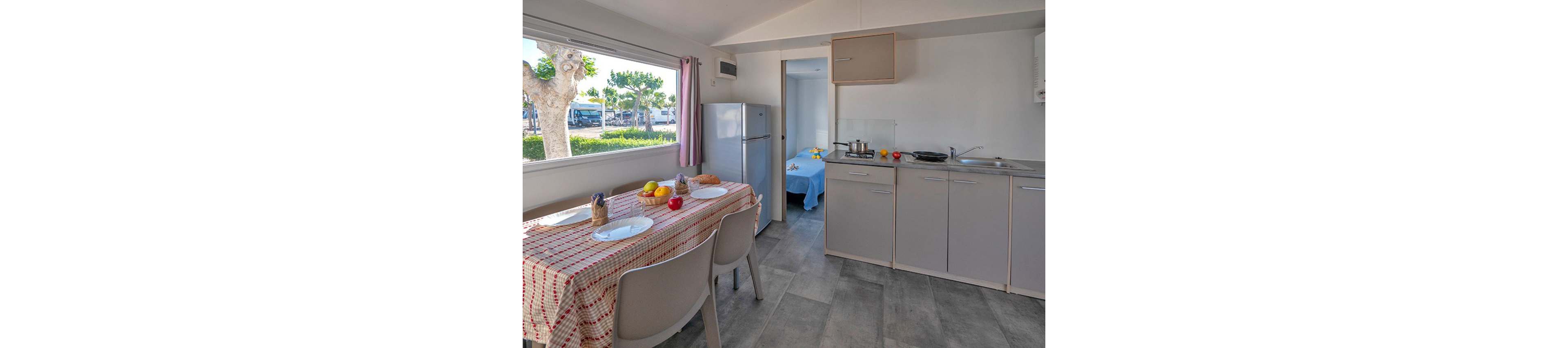A small modern kitchen with grey cabinets and a refrigerator opens into a cozy dining area. A table covered with a red and white checkered cloth is set with plates, fruit, and bread, suggesting a casual meal. Through an open doorway, a neatly made bed with a blue cover is visible, indicating a studio or holiday accommodation. Large windows bring in natural light and show greenery and parked vehicles outside, creating a relaxed vacation atmosphere.