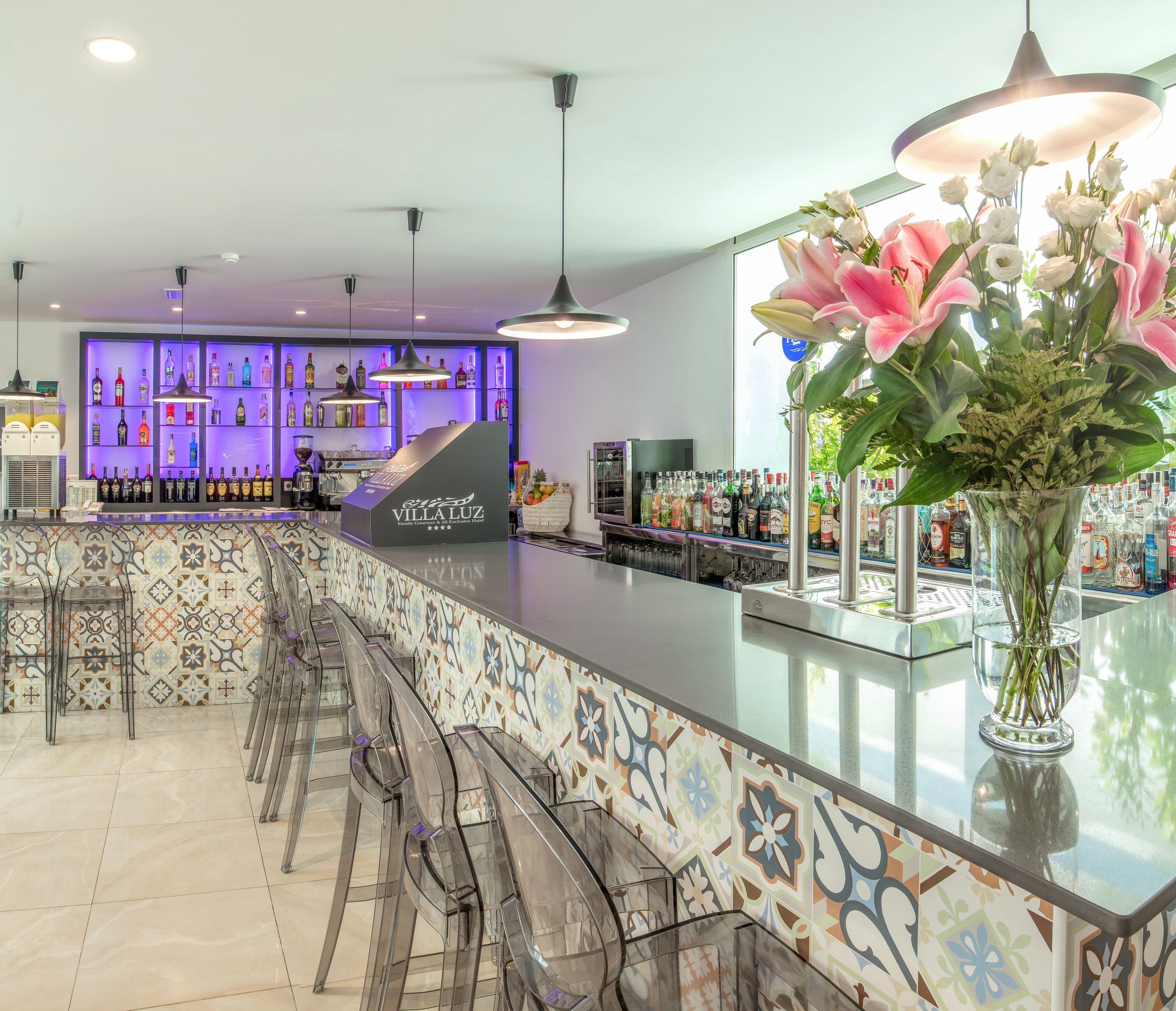 Bright contemporary bar interior featuring a long counter with colorful patterned tiles and transparent bar stools. The setting includes a backlit shelving unit displaying various bottles and glassware, creating a vibrant nightlife atmosphere. Pendant lights hang from the ceiling, illuminating the glossy countertop and reflective surfaces. A tall glass vase with pink lilies and white flowers adds a fresh decorative touch to the bar area.