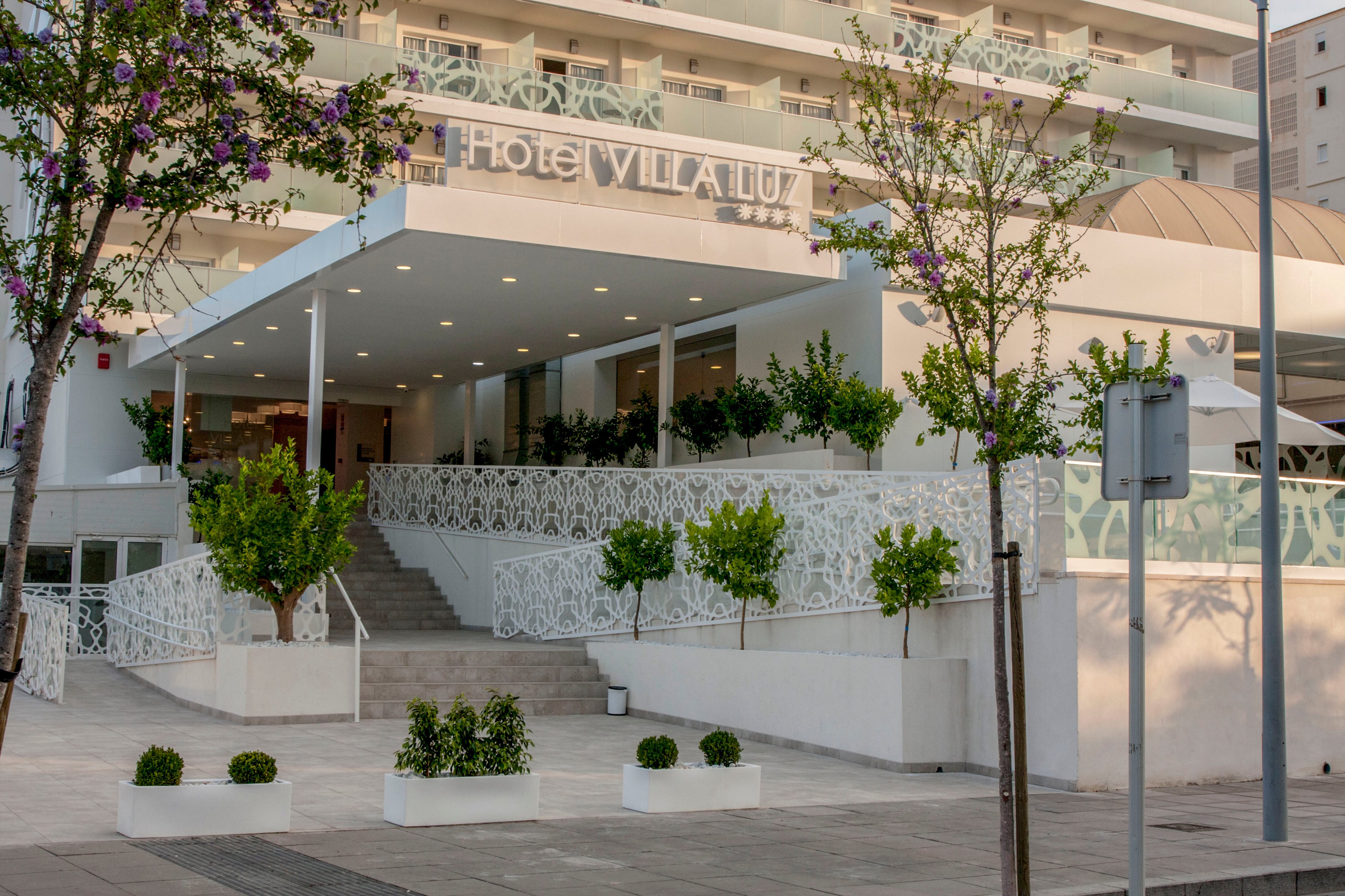 Contemporary exterior view of Hotel Villa Luz shows a bright, welcoming entrance. A wide staircase and ramp with decorative white railings lead up to the main lobby under a flat canopy. Potted shrubs and small trees line the pathway, adding greenery to the clean, minimalist architecture. The multi-story building facade and nearby street elements suggest an urban or coastal tourist setting.