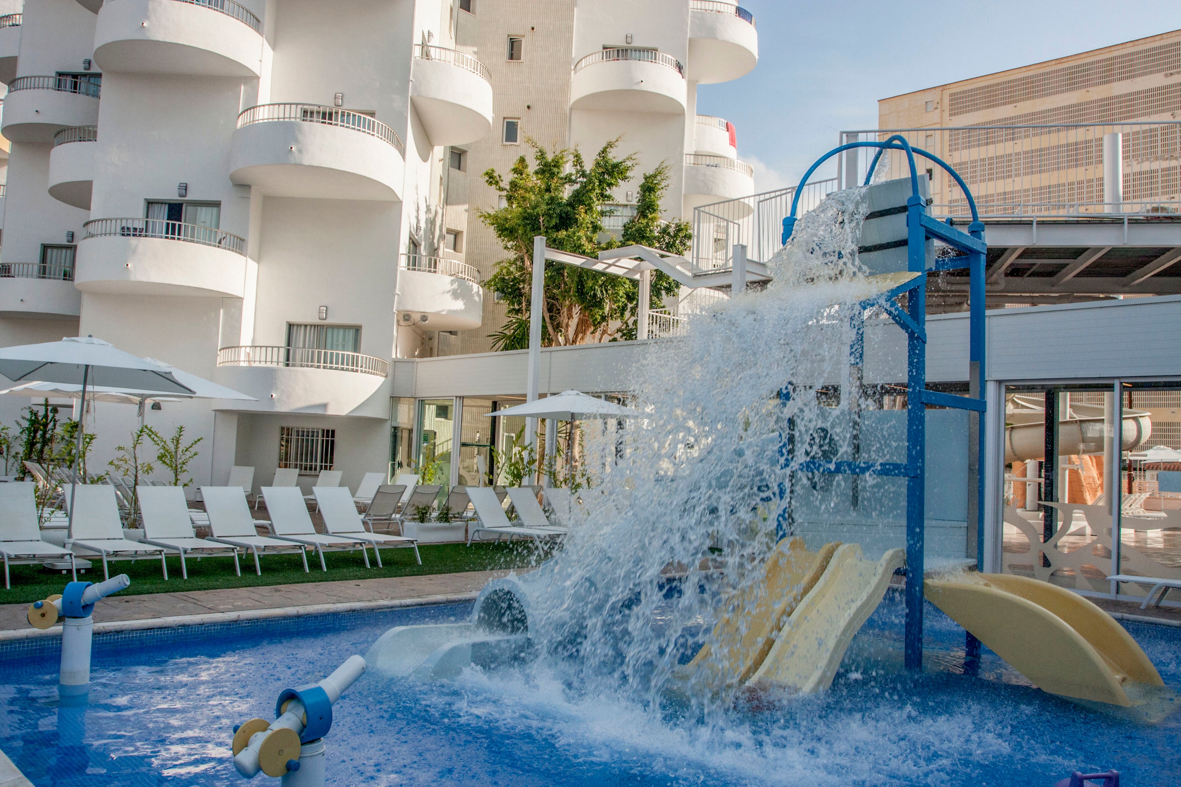 A small water play area with slides and a splash feature is set in the courtyard of a modern hotel. White lounge chairs line the poolside, facing the blue shallow pool and water jets. Curved balconies and white building facades rise in the background, with some greenery adding contrast. The scene suggests a family-friendly resort environment in warm daylight.