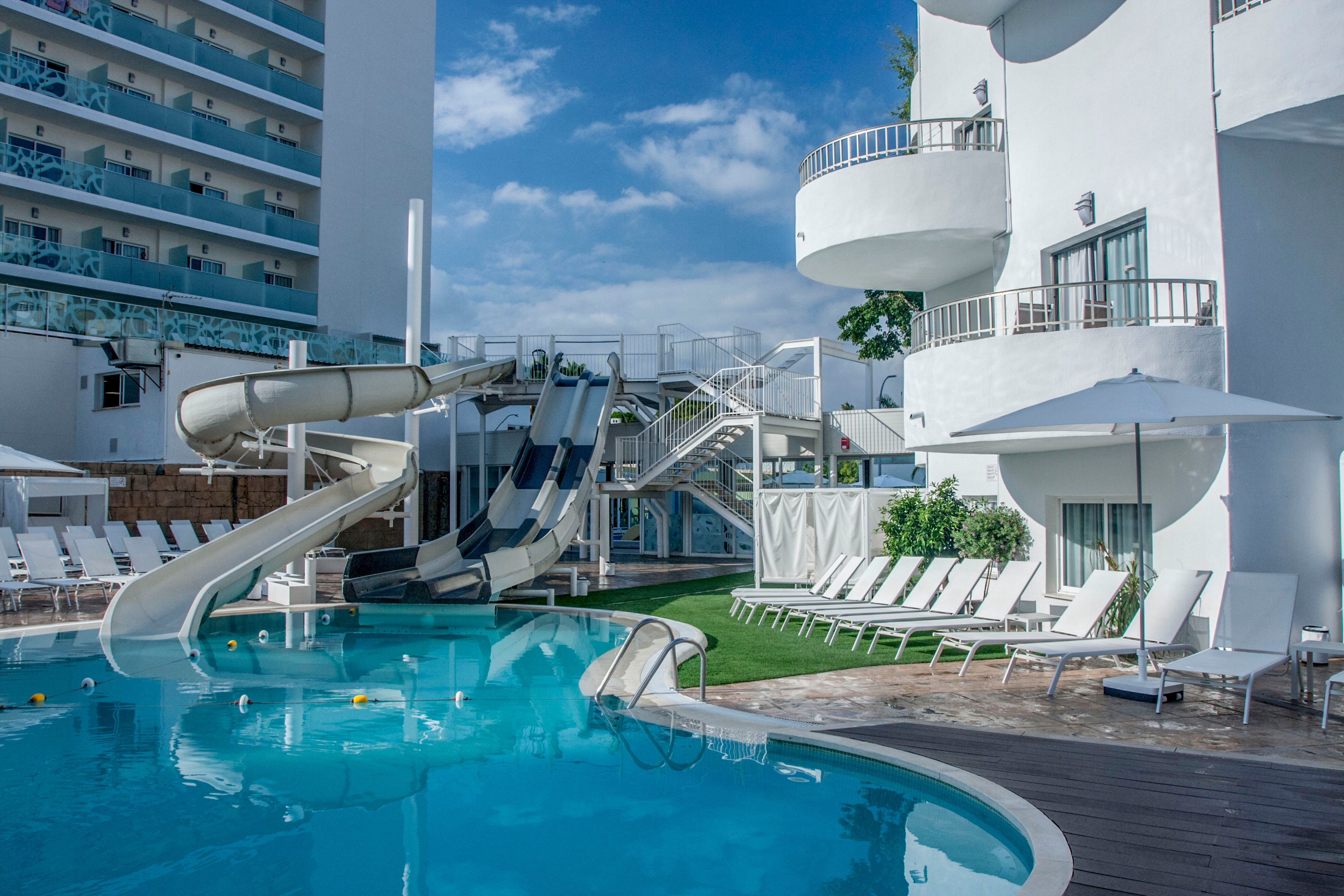 A bright outdoor hotel pool area features curved white waterslides leading into a clear blue swimming pool. White lounge chairs line the poolside deck under large umbrellas, creating a relaxed vacation setting. Contemporary white buildings and balconies surround the pool, with railings and walkways connecting different levels. The scene is captured on a sunny day with blue sky and scattered clouds, emphasizing a clean, modern resort atmosphere.