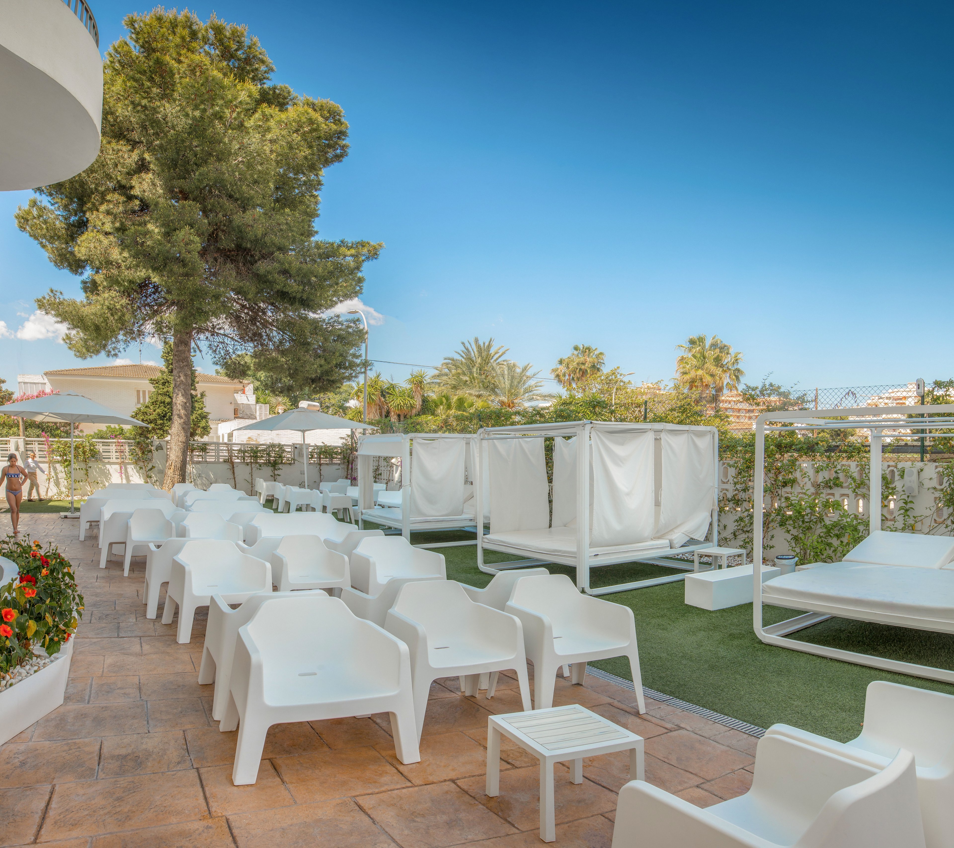 An outdoor resort lounge area features rows of modern white plastic chairs and small tables on a tiled terrace. To the right, covered daybeds and green artificial grass create a relaxed sunbathing zone. Tall trees, lush plants, and distant buildings frame the space under a clear blue sky. The scene suggests a tranquil vacation setting designed for comfort and leisure.