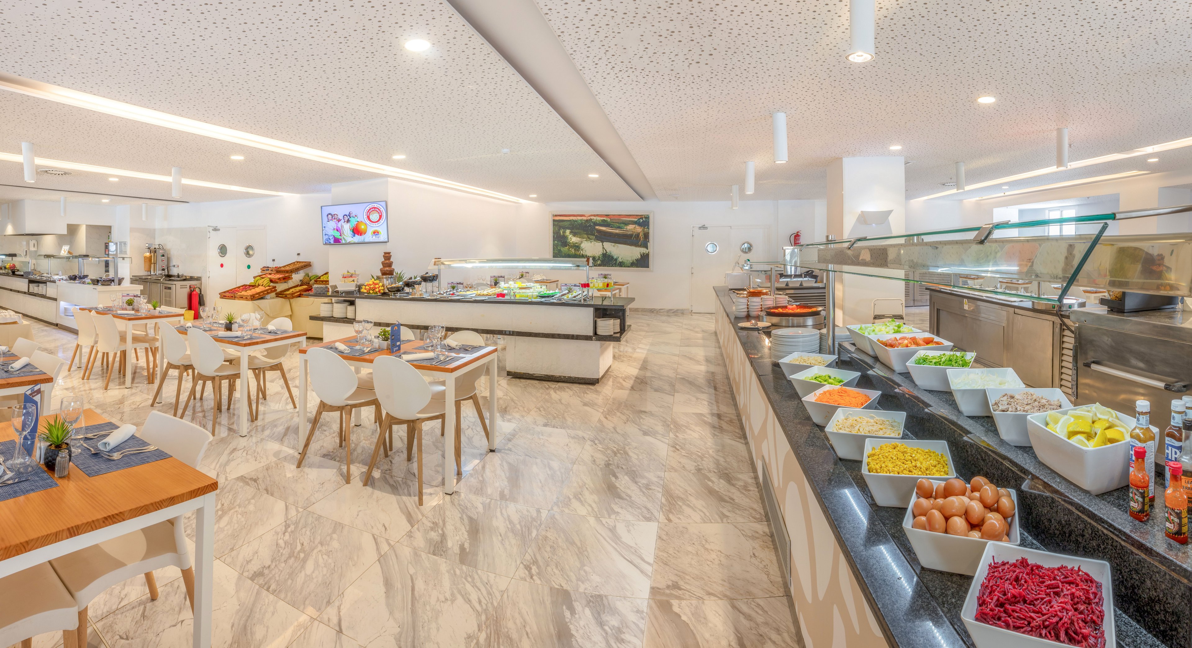 A spacious self-service restaurant features a long buffet counter filled with salads, vegetables, eggs, and other cold dishes. Modern white chairs and wooden tables are neatly arranged on a polished marble floor, creating an airy dining area. The ceiling has recessed lighting and acoustic panels, while glass sneeze guards protect the food stations. A digital screen and wall art add contemporary touches to the clean, organized interior.