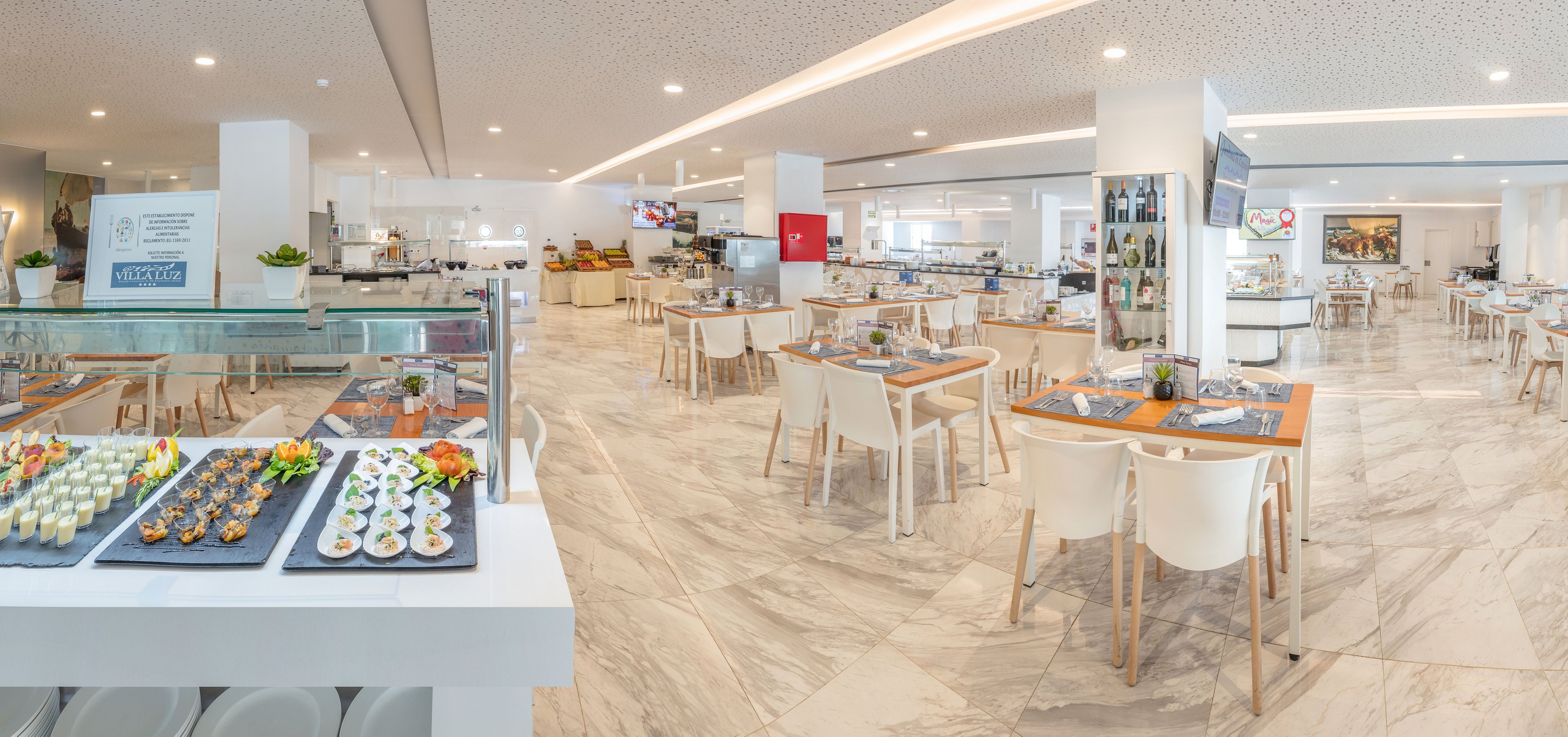 Panoramic view of a spacious, contemporary buffet restaurant with neatly arranged tables and chairs. The foreground shows a glass display counter featuring assorted appetizers and desserts on trays. In the background, multiple food stations, drink areas, and wall-mounted screens are visible, all set within a clean, white-toned environment. The light marble flooring and minimalist decor create an airy, inviting dining atmosphere.