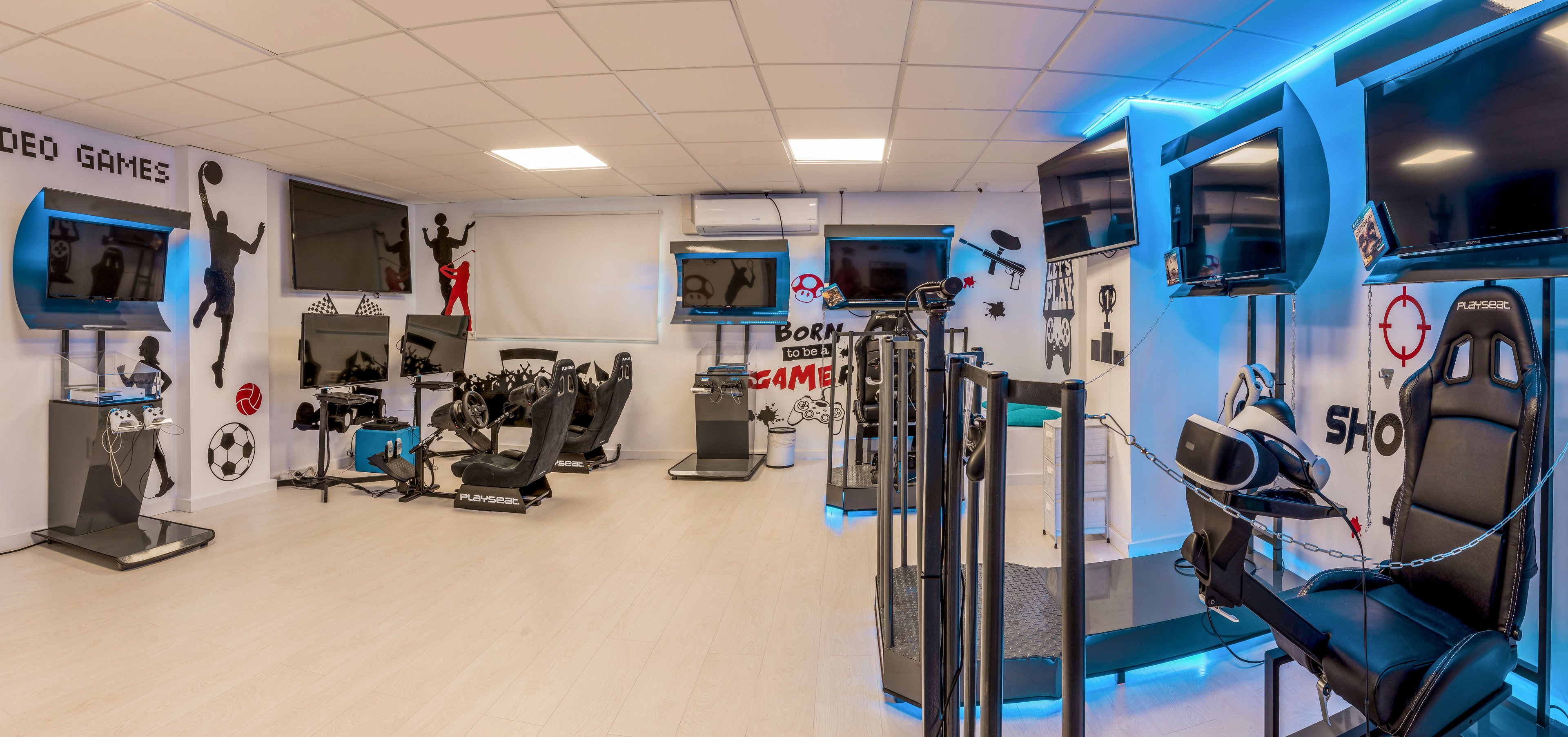 Panoramic view of a spacious gaming room featuring multiple large wall-mounted screens and specialized gaming stations. Racing simulators with steering wheels and pedals are positioned alongside a virtual reality setup with headsets and motion platforms. The walls are decorated with dynamic gamer silhouettes and text such as "VIDEO GAMES" and "GAMES" that emphasize the entertainment theme. Cool blue accent lighting and sleek black equipment create a futuristic, high-tech atmosphere.