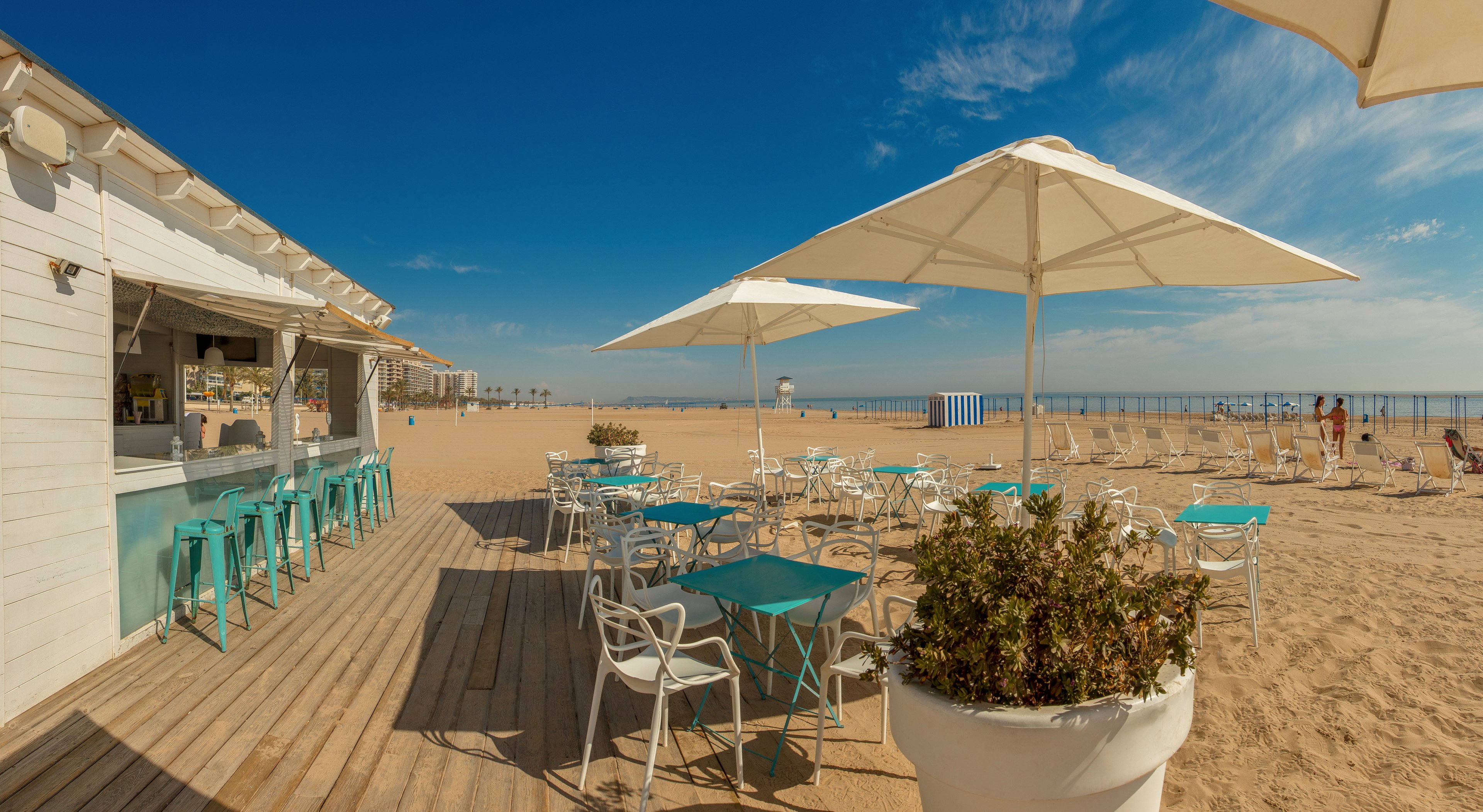 An open-air beach bar with turquoise stools and white tables sits on a wooden deck beside a sandy shoreline. Large white umbrellas provide shade over the seating area, while the expansive beach stretches toward the calm sea. A clear blue sky with scattered clouds enhances the bright, summery atmosphere. In the distance, beach chairs and a striped changing cabin add to the relaxed coastal setting.