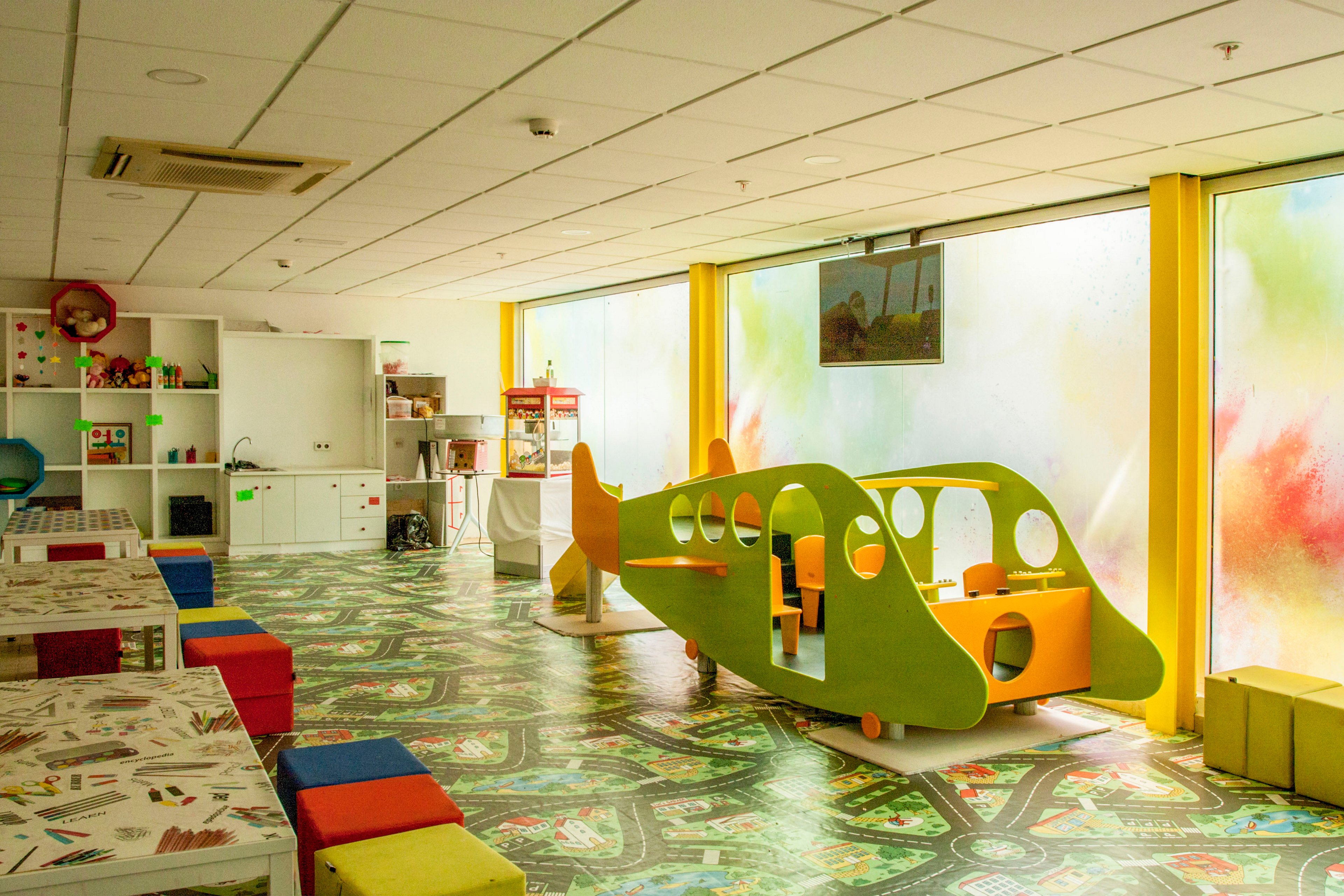 Bright children’s playroom featuring a green and yellow airplane-shaped play structure stands in the center of the room. Long tables with art supplies and multicolored cube seats line one side, while shelves with toys and storage units are visible in the background. Large windows with colorful abstract patterns let in natural light, creating a cheerful atmosphere. The patterned floor and vivid primary colors emphasize a fun, creative environment for kids.