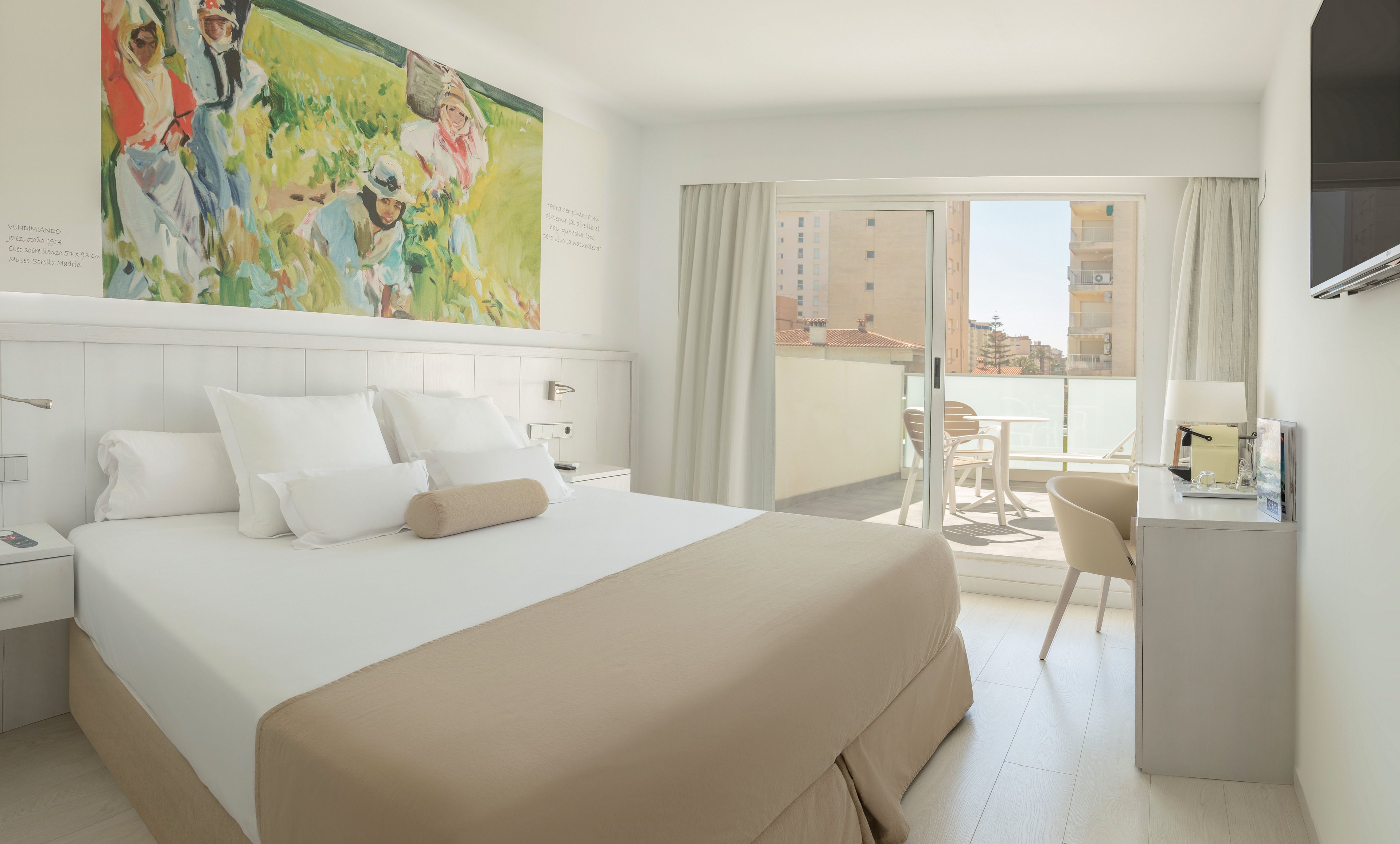 A contemporary hotel room features a large neatly made bed with white linens and beige accents. Above the headboard hangs a colorful painting depicting people in a field. Sunlight floods the space through sliding glass doors that open onto a private terrace with outdoor seating. A desk and chair sit beside the window, and a wall-mounted television is visible on the right, creating a clean, comfortable atmosphere.