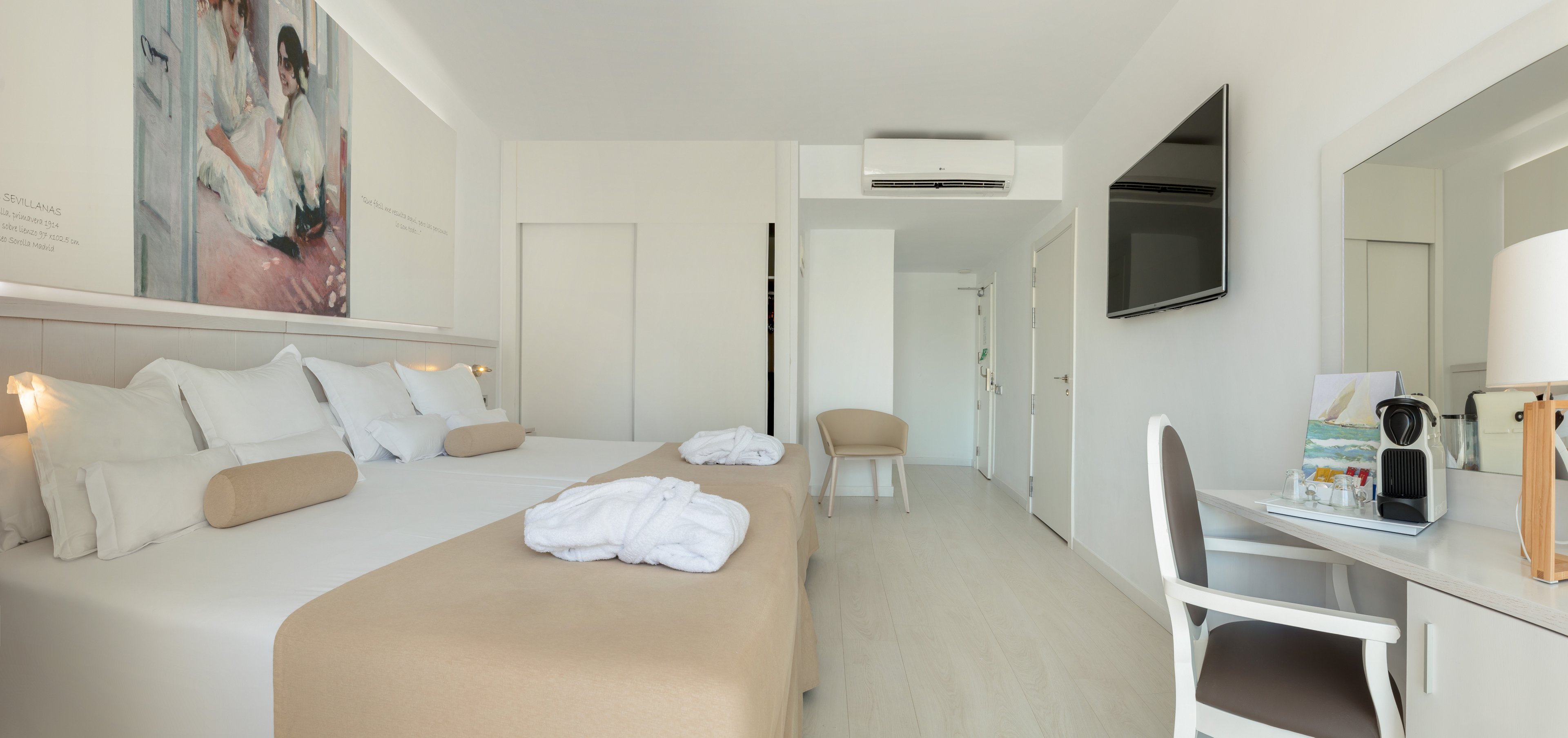 A spacious, contemporary hotel room features a large bed with white linens and beige accents. A desk with a chair, coffee machine, and decorative items sits opposite a wall-mounted flat-screen television. The room is decorated in a light, neutral palette with minimalist furniture and a soft, airy atmosphere. A wall art piece above the bed and neatly placed bathrobes add a touch of comfort and style.