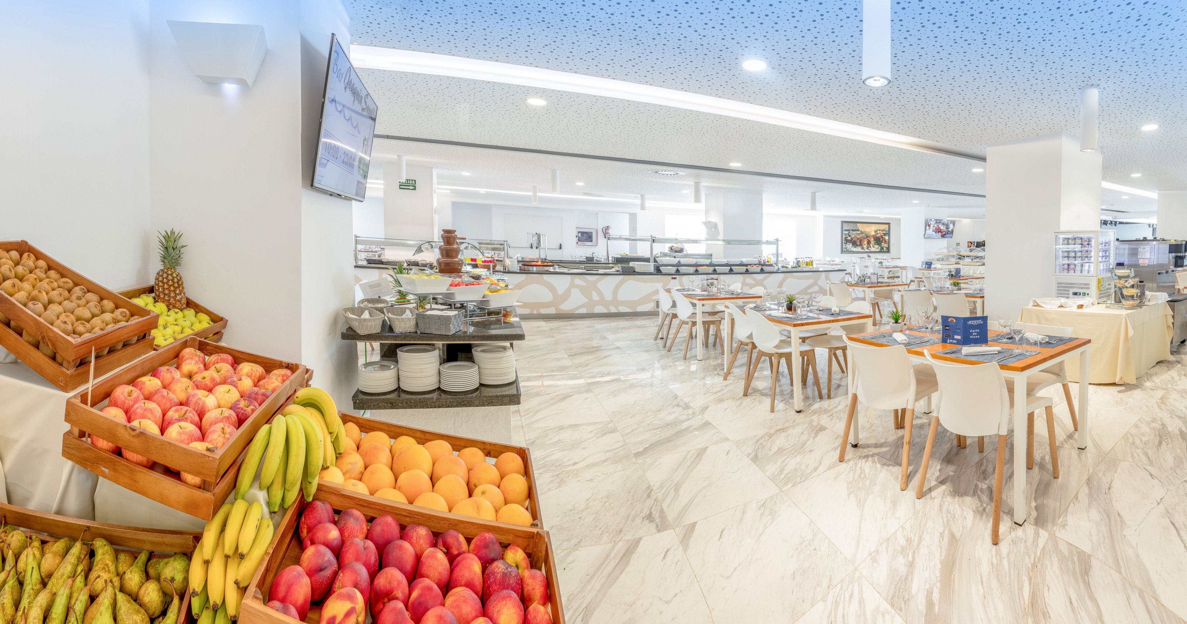 A spacious self-service restaurant features neatly arranged wooden crates filled with bananas, oranges, and other fresh fruits in the foreground. The interior is bright and modern, with white walls, sleek buffet counters, and marble-style flooring. Several white tables and chairs are set for guests, and serving stations line the back wall. The overall atmosphere is clean, inviting, and designed for casual dining.