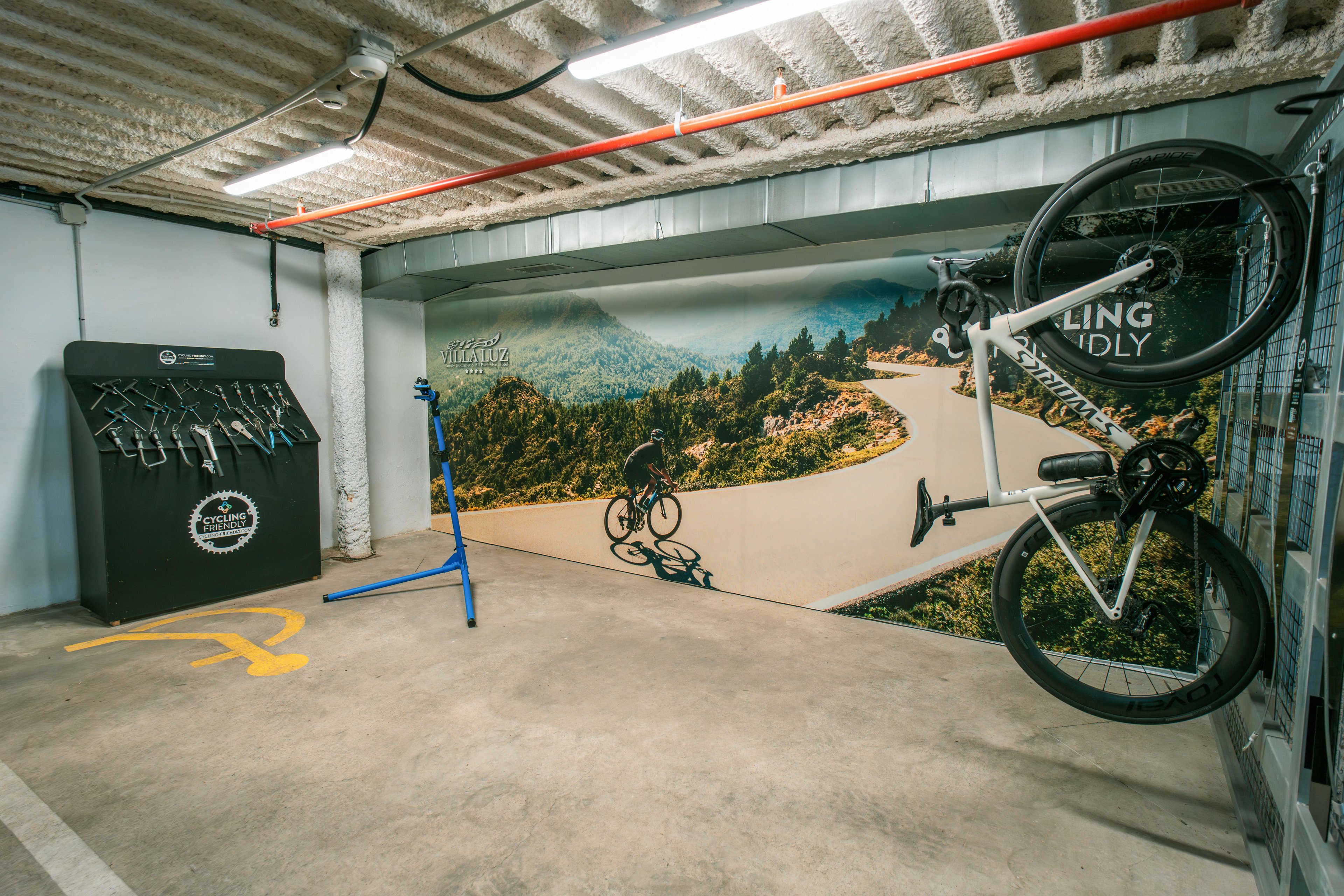 An indoor bike storage room features a wall-mounted bicycle and a blue repair stand in the center. A large mural on the back wall shows a cyclist riding along a winding mountain road. On the left, a black equipment station holds multiple hanging tools and accessories. The concrete floor and exposed ceiling pipes give the space an industrial look, with a yellow bike symbol painted on the ground.