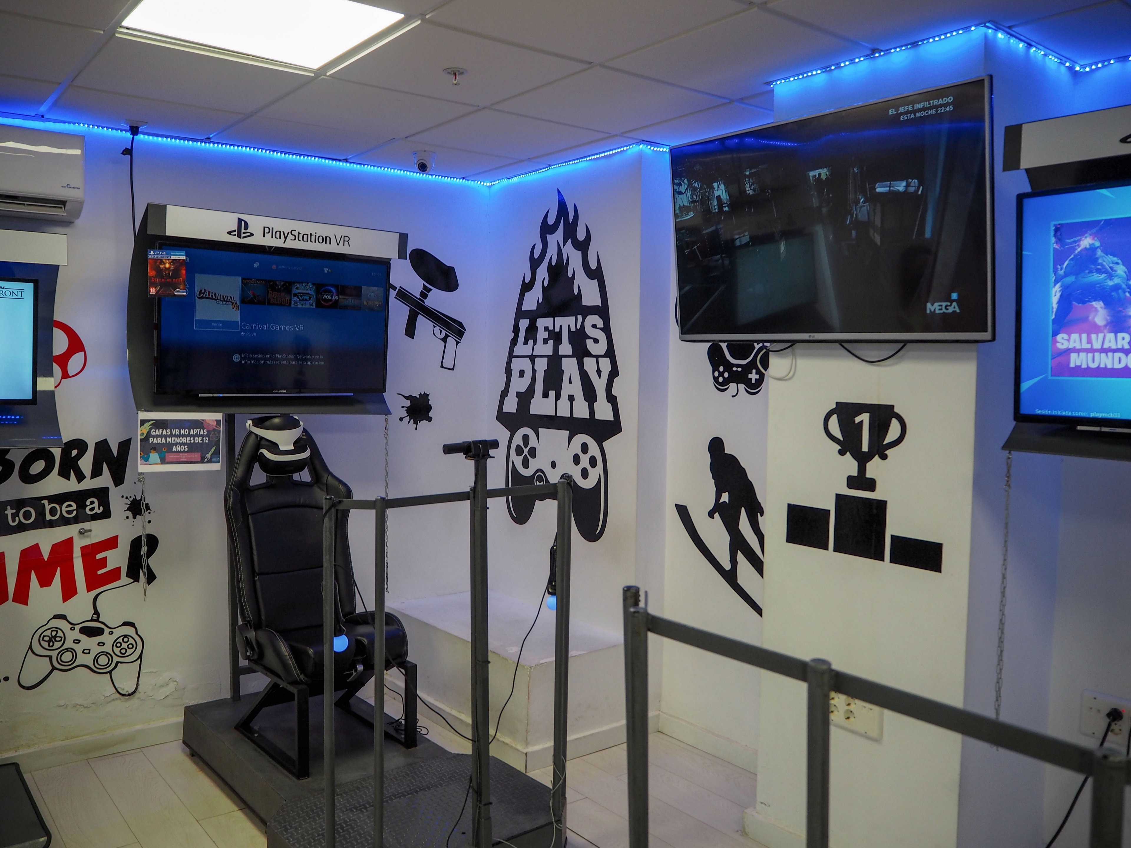 A dedicated PlayStation VR gaming station is set up in a brightly lit arcade-style room. A racing-style chair and VR sensors face a wall-mounted screen labeled PlayStation VR, surrounded by gaming-themed wall art. Blue LED strip lighting accents the ceiling and walls, and additional monitors display gameplay and promotional content. The space is designed for immersive virtual reality experiences and social gaming.