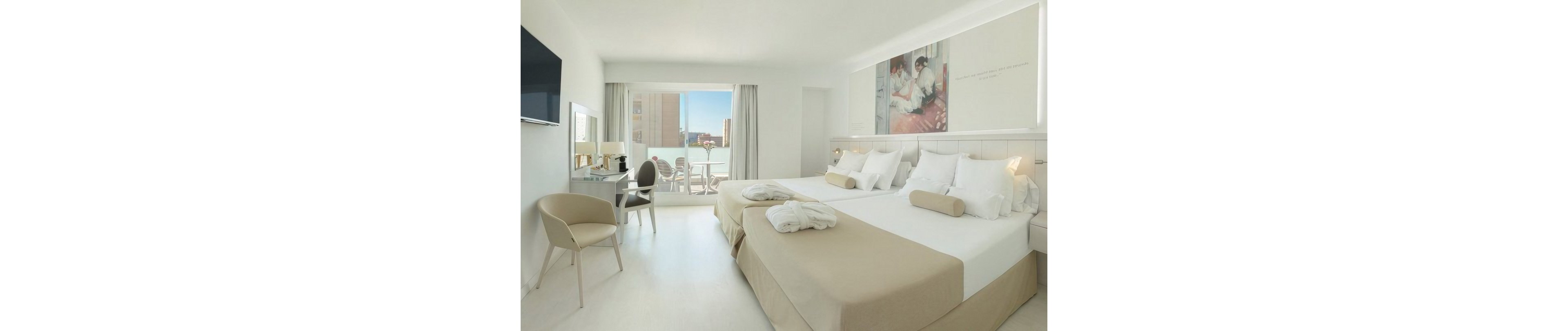 A spacious, contemporary hotel room features two large beds with white linens and beige accents. The space is decorated in a minimalist style, with a wall-mounted television, a small desk area, and a light-colored chair. Floor-to-ceiling curtains frame a balcony doorway that opens to an outdoor seating area and city buildings in the background. Soft natural light and neutral tones create a calm, inviting atmosphere.