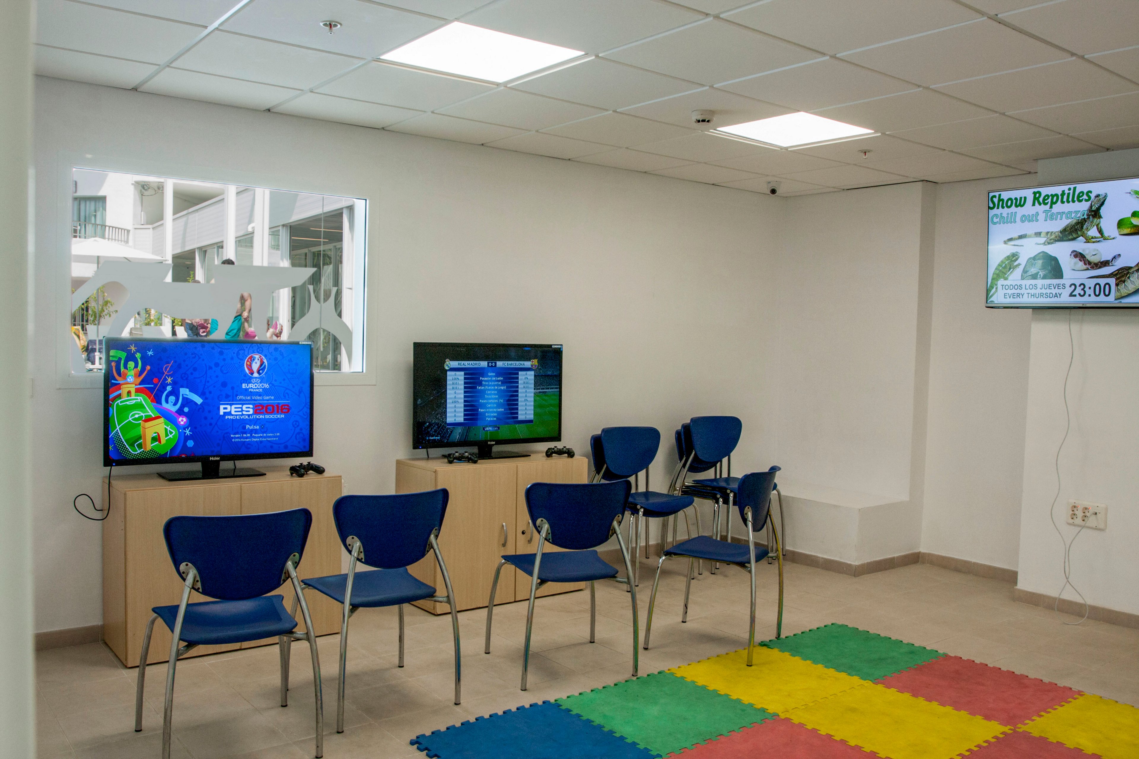 A bright waiting room features two televisions on a wooden cabinet, each connected to gaming consoles with controllers. Several blue chairs are arranged in front of the screens, suggesting a casual play area or entertainment zone. A colorful foam mat with red, yellow, green, and blue squares lies on the floor, adding a playful touch. On the wall, a digital display advertises a reptile show with the time clearly shown as 23:00.