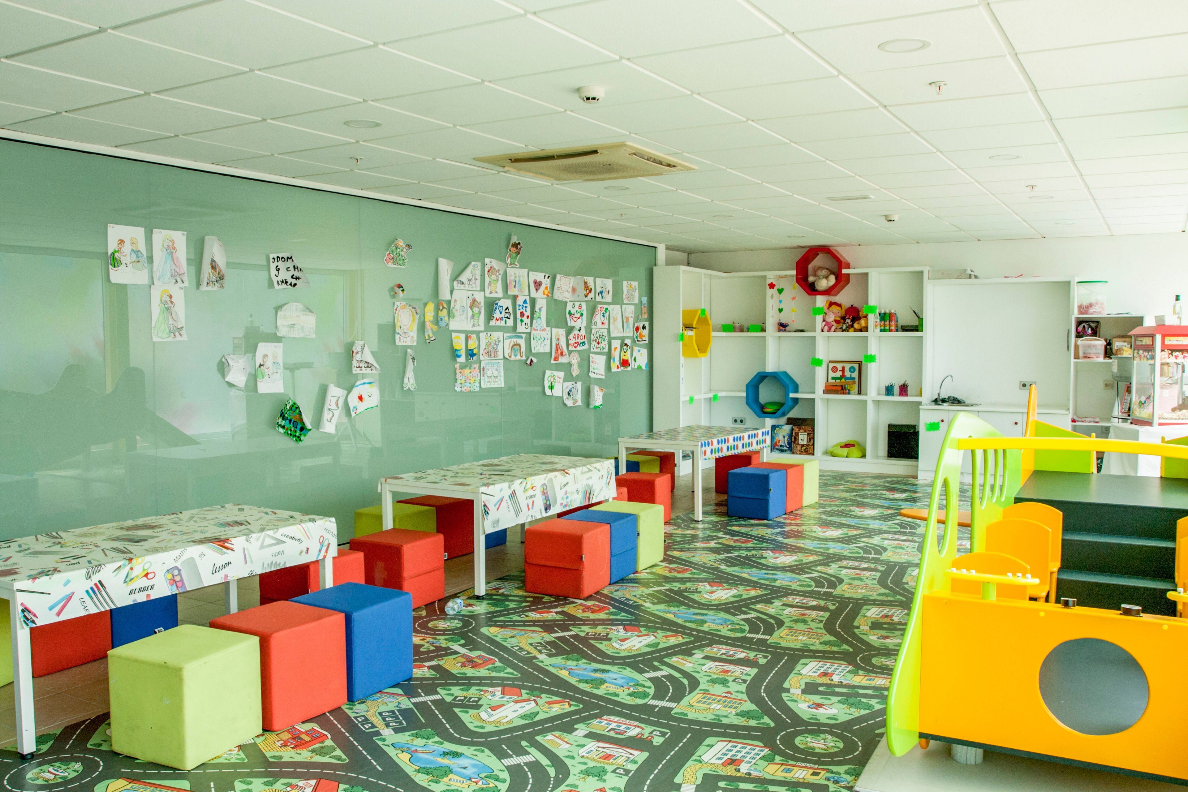 Spacious children’s playroom featuring low tables and multicolored cube seats. The room has a patterned floor with road and city graphics and a large glass wall displaying kids’ drawings. Open shelving holds toys and play equipment, while a yellow and green play structure stands to the right. The overall atmosphere is cheerful, creative, and designed for early childhood activities.