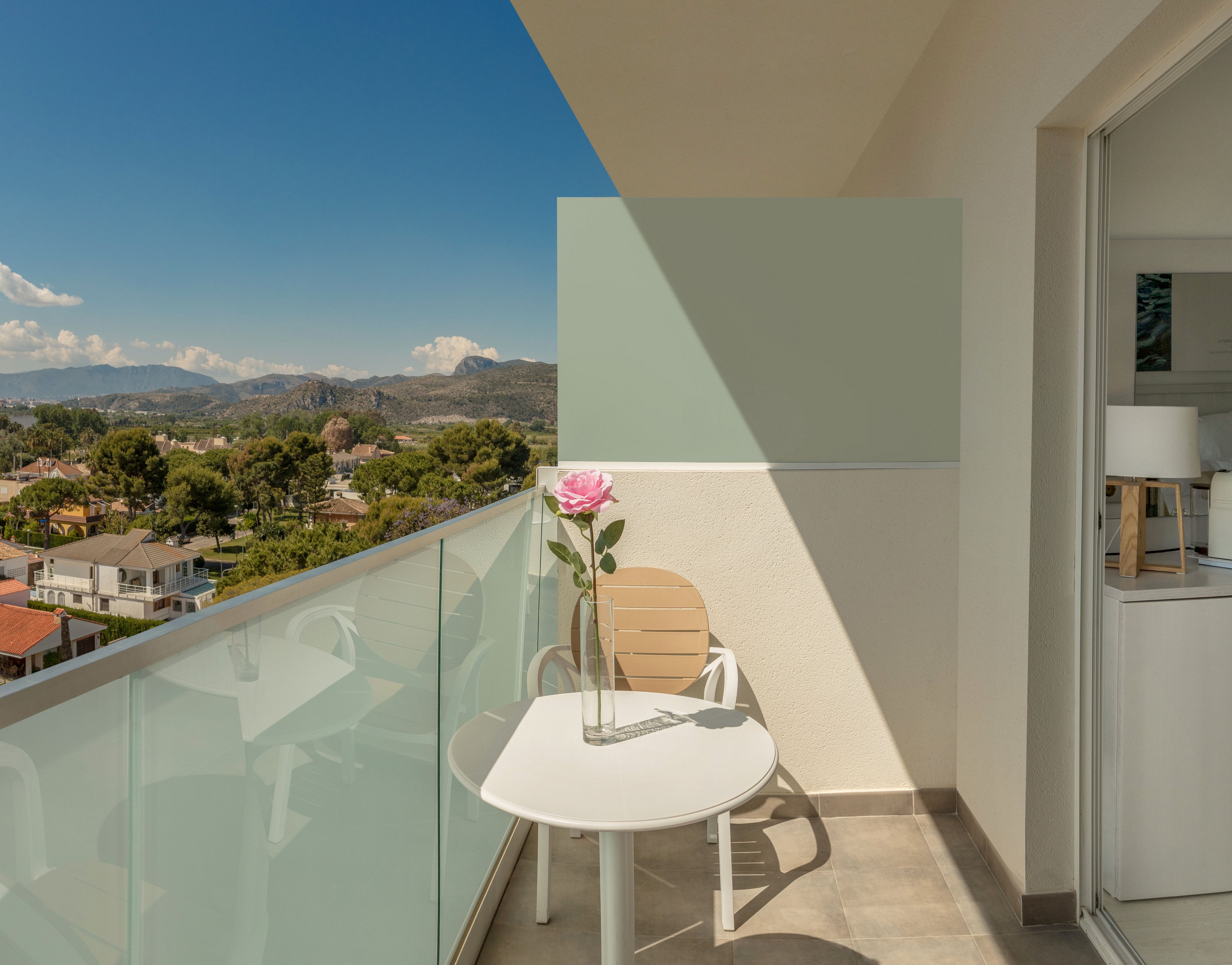 A bright modern balcony features a small round white table with a single pink flower in a clear glass vase. A light-colored chair sits beside the table, facing outward toward a scenic residential landscape and distant mountains. Glass railing panels frame the balcony, while soft neutral walls and a partially visible interior with a lamp create a calm, contemporary atmosphere. Clear blue sky and natural greenery enhance the feeling of relaxation and openness.