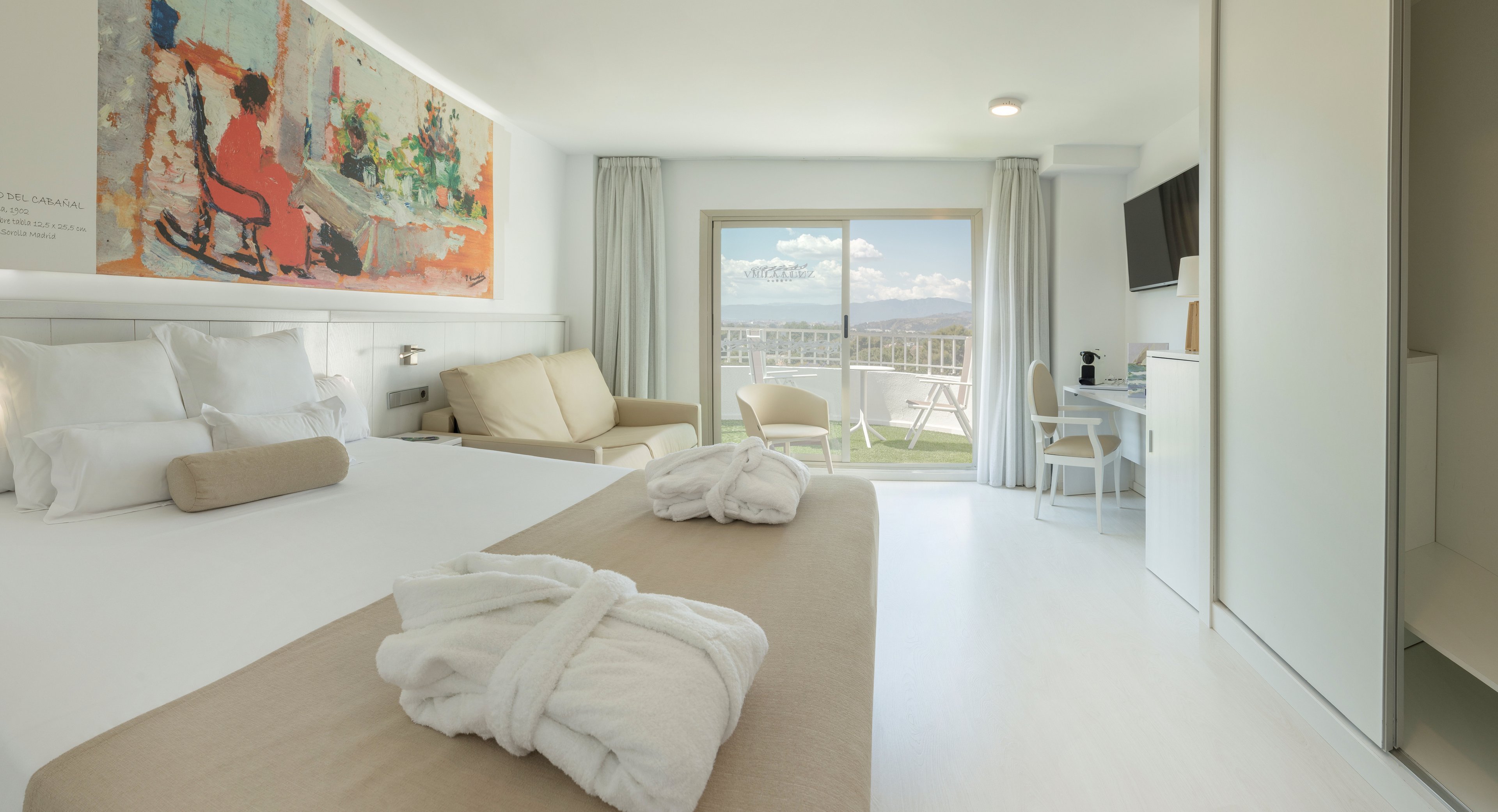 Spacious contemporary hotel room featuring a large bed with beige accents and neatly folded bathrobes. A sofa seating area and wall-mounted television face a sliding glass door that opens to a balcony. Soft neutral tones and white furnishings create a clean, minimalist atmosphere, complemented by a colorful artwork above the bed. The balcony offers outdoor chairs and a scenic view of distant mountains under a bright sky.