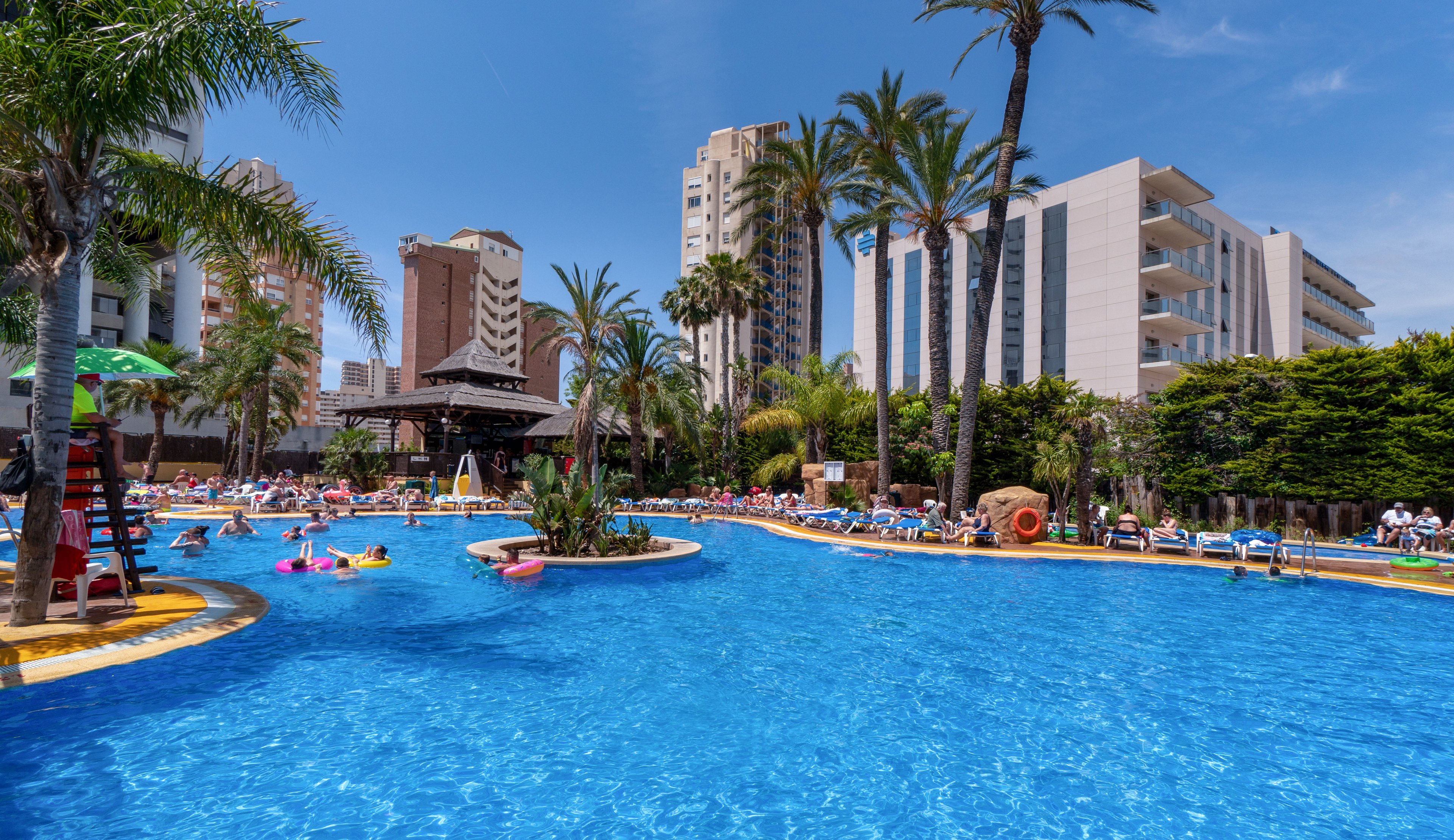 A large outdoor swimming pool is filled with people relaxing on inflatables and sun loungers. Tall palm trees line the poolside, adding a tropical feel to the urban resort setting. Modern highrise apartment and hotel buildings rise in the background under a clear blue sky. The scene suggests a busy vacation atmosphere with families and tourists enjoying the sunny weather.