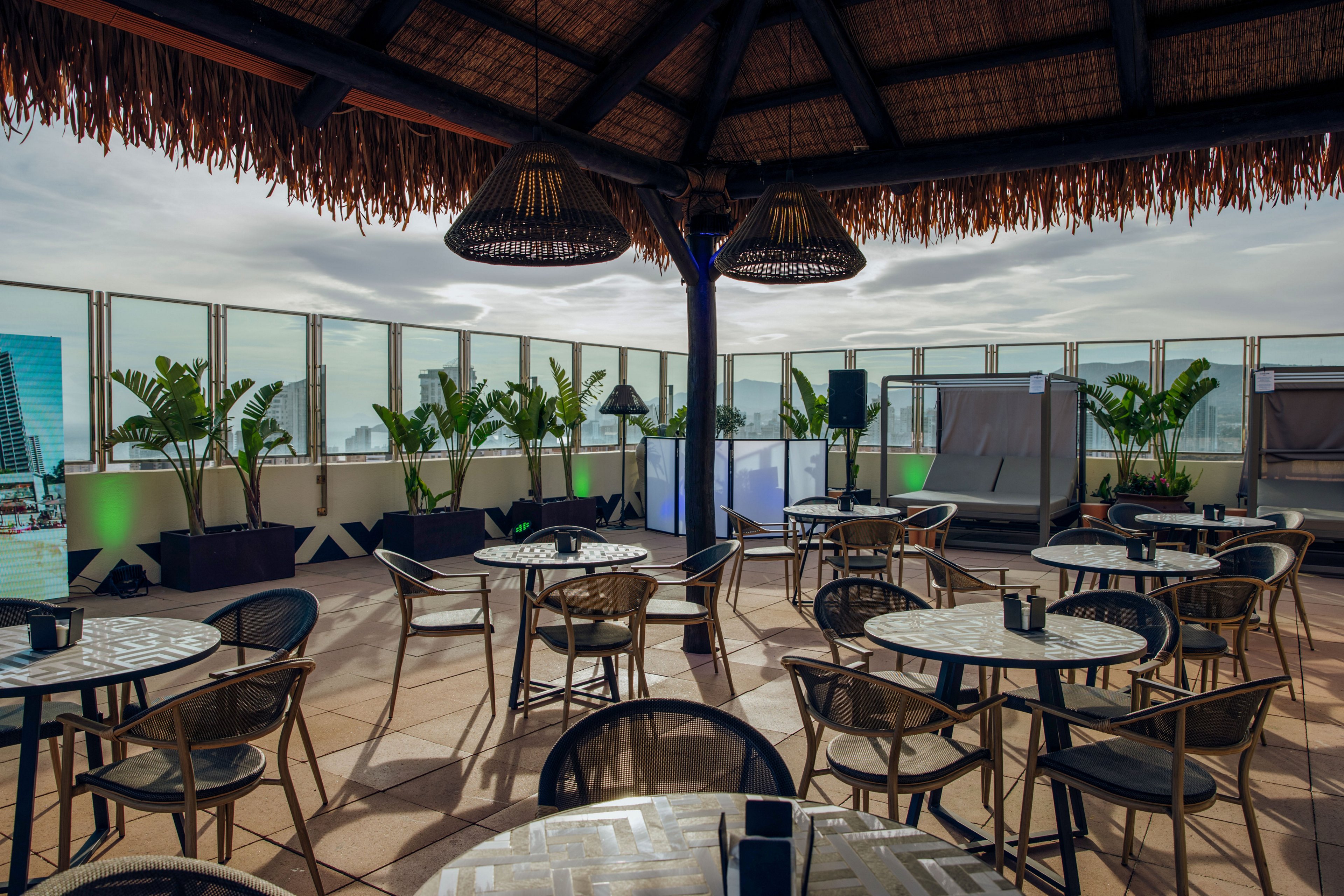 An open-air rooftop terrace features multiple round tables and chairs arranged for casual seating. The space is covered by a thatched roof structure and surrounded by glass railings, offering views of nearby city buildings. Potted tropical plants line the perimeter, adding greenery to the modern setting. Soft lighting and a small bar or DJ area suggest a relaxed social or event atmosphere.
