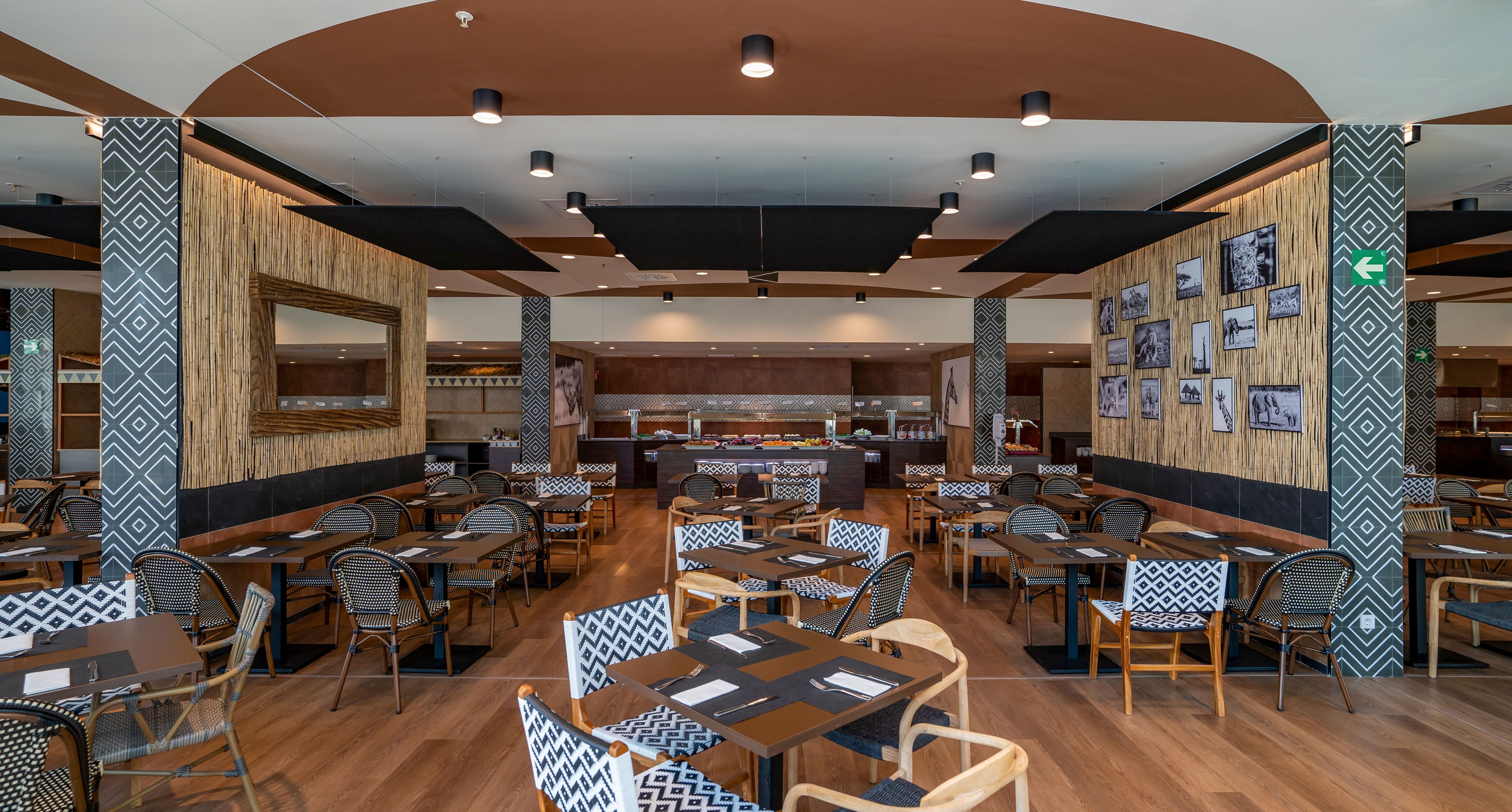 Spacious restaurant dining area with neatly arranged tables and chairs is shown empty and ready for guests. The setting features warm wooden flooring, patterned columns, and decorative wall panels with framed black-and-white photos. A large mirror and ceiling lights add brightness, while the buffet counter is visible in the background. The overall atmosphere feels contemporary, organized, and welcoming.