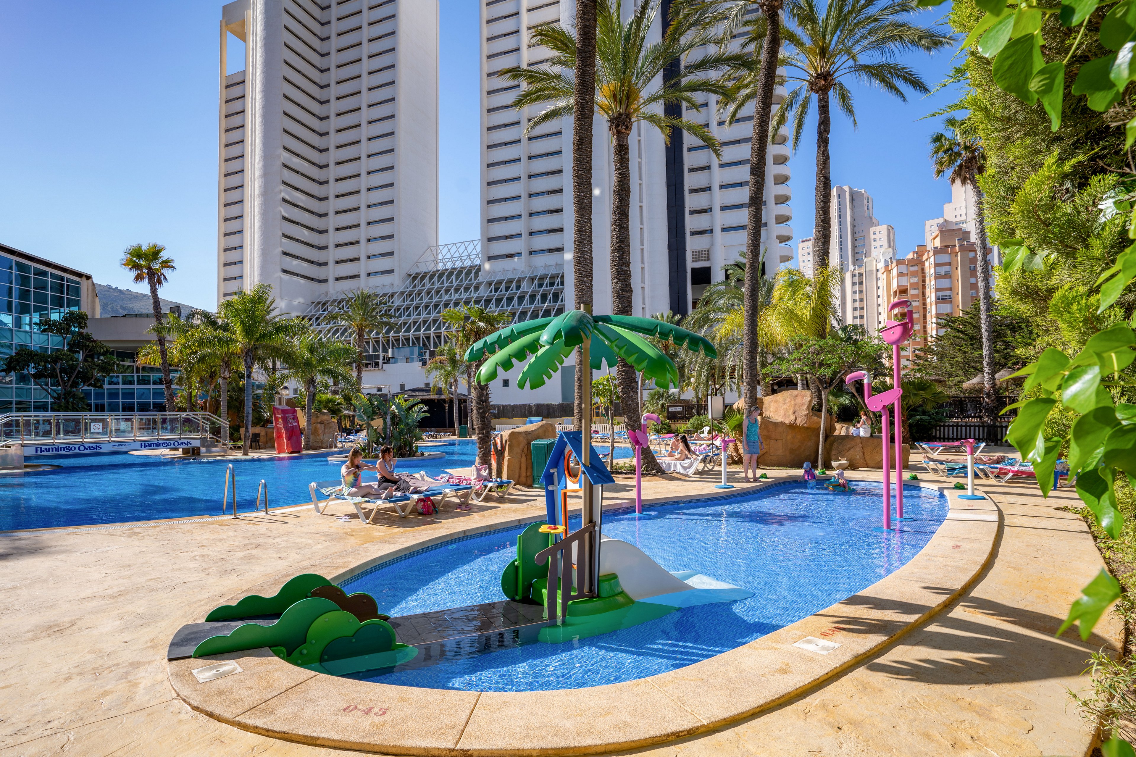 A bright resort swimming pool features colorful children’s water play structures, including palm tree sprayers and slides. Tall modern high-rise buildings rise in the background, framed by lush palm trees and tropical landscaping. Sun loungers and umbrellas line the poolside area, where guests relax under clear blue skies. The overall scene conveys a fun, family-friendly vacation atmosphere in a warm, sunny destination.