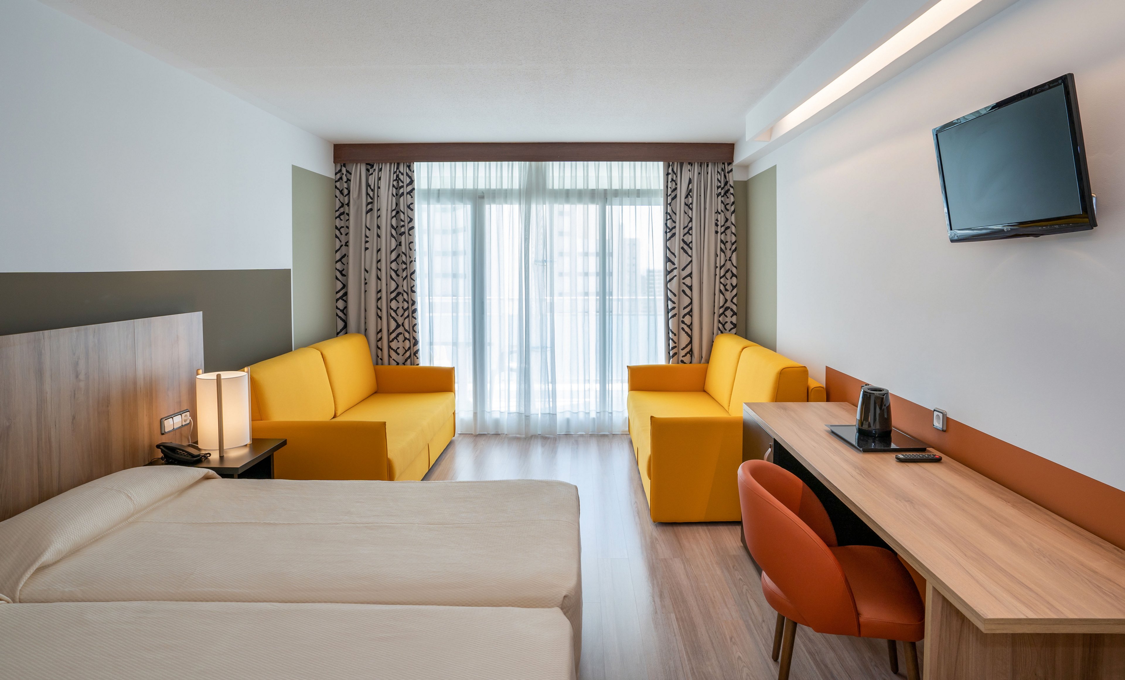 Contemporary hotel room interior featuring two neatly made twin beds in the foreground. The space includes a long wooden desk with an orange chair, telephone, and electric kettle, facing a wall-mounted television. Two bright yellow sofas sit near large floor-to-ceiling windows with sheer curtains and patterned drapes, allowing natural light to fill the room. The overall design is clean and minimalist with warm wood tones and bold accent colors.