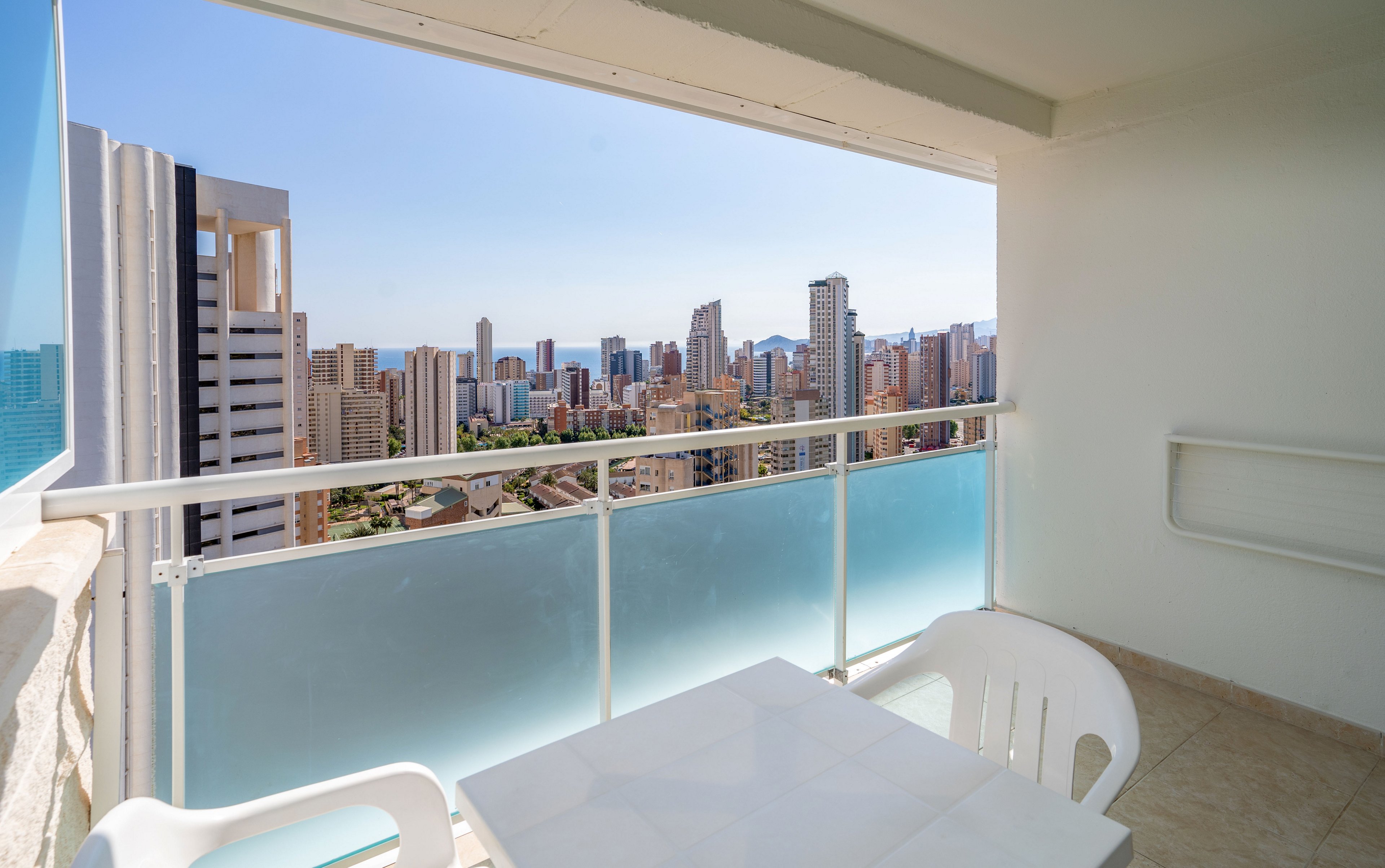 A bright apartment balcony overlooks a modern city skyline filled with tall buildings. The foreground shows a white plastic table and chairs on a tiled floor beside a frosted glass railing. Clear blue sky and distant high-rises create an open, airy atmosphere. The scene suggests a relaxing urban outdoor space with panoramic views.
