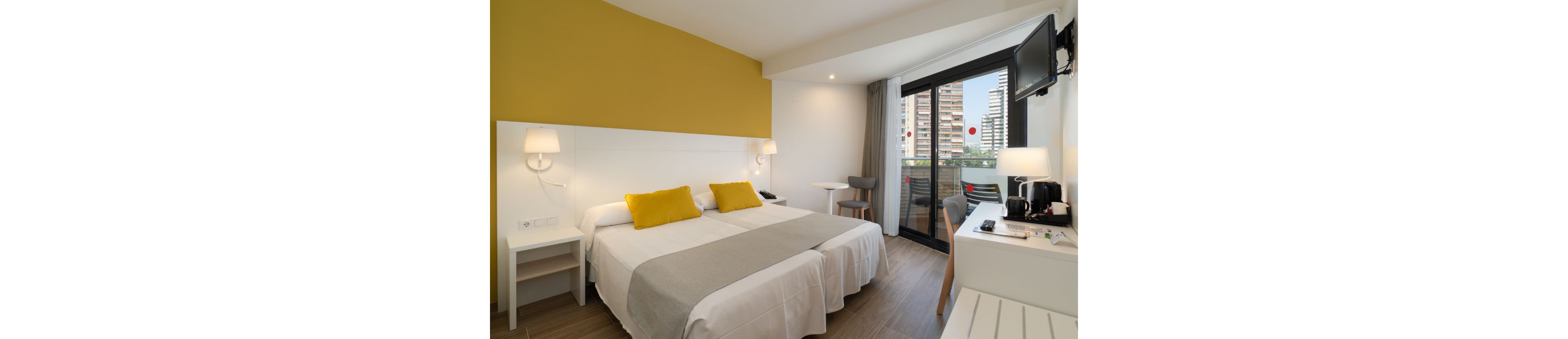 Bright contemporary hotel room featuring two single beds with white linens and gray runners. The space includes a yellow accent wall, bedside lamps, and a small desk area with a chair. A wall-mounted television and a refreshment station with a kettle sit opposite the beds. Large glass doors open to a balcony overlooking tall city buildings and outdoor seating.