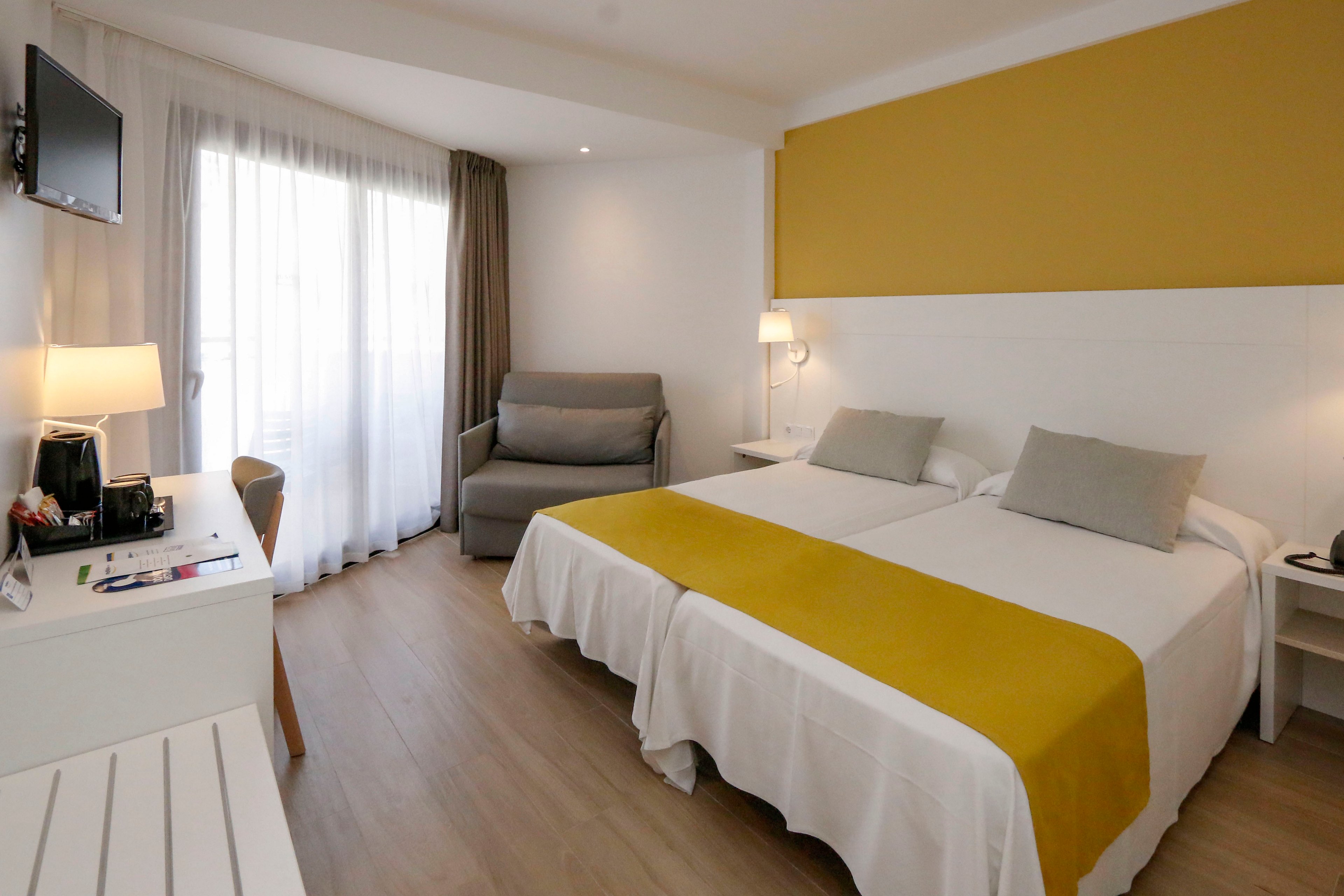 A contemporary hotel room features two neatly made twin beds with white linens and mustard yellow runners. The space includes a gray armchair near a large window with sheer and blackout curtains, allowing natural light to fill the room. A wall-mounted television, bedside tables with lamps, and a small desk area with amenities create a comfortable, functional environment. The overall design is minimal and clean, with warm tones and wood flooring contributing to a cozy atmosphere.