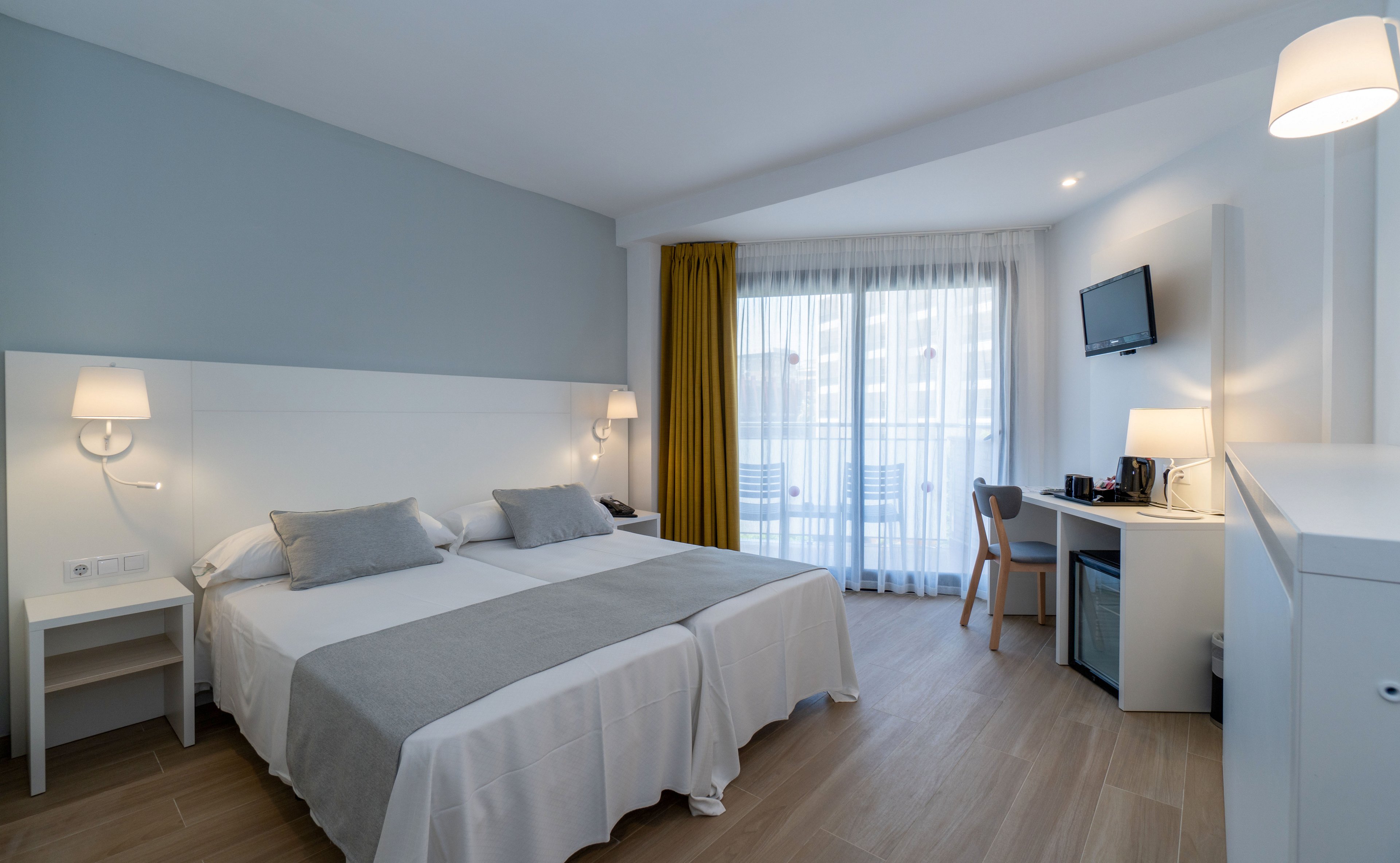 Bright contemporary hotel room featuring two twin beds with white linens and gray throws. A small desk area with a wall-mounted television, coffee station, and mini fridge sits to the right. Floor-to-ceiling windows with sheer curtains and a yellow blackout drape open to a balcony with outdoor seating. Soft wall sconces and neutral tones create a clean, minimalist atmosphere.