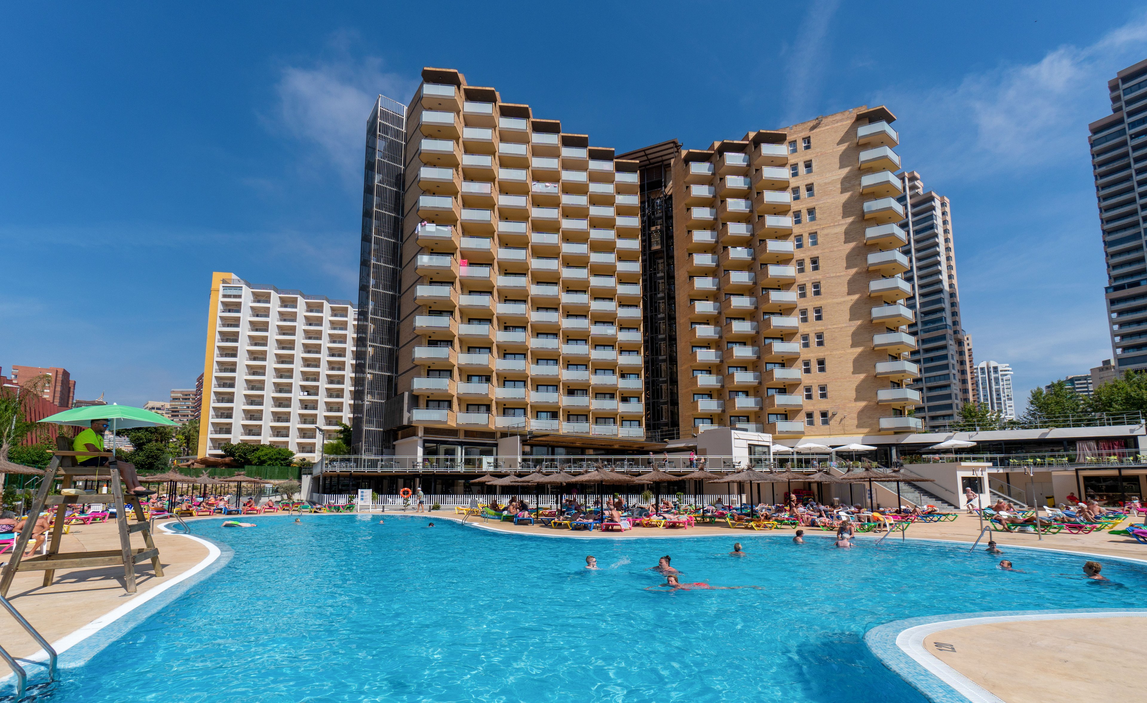 A large outdoor swimming pool is filled with people relaxing and playing in the water. Tall multi-story hotel buildings rise in the background, with balconies facing the pool area. Sun loungers and umbrellas line the poolside, creating a busy holiday atmosphere. A lifeguard sits on a raised chair, overseeing the scene on a bright, sunny day.