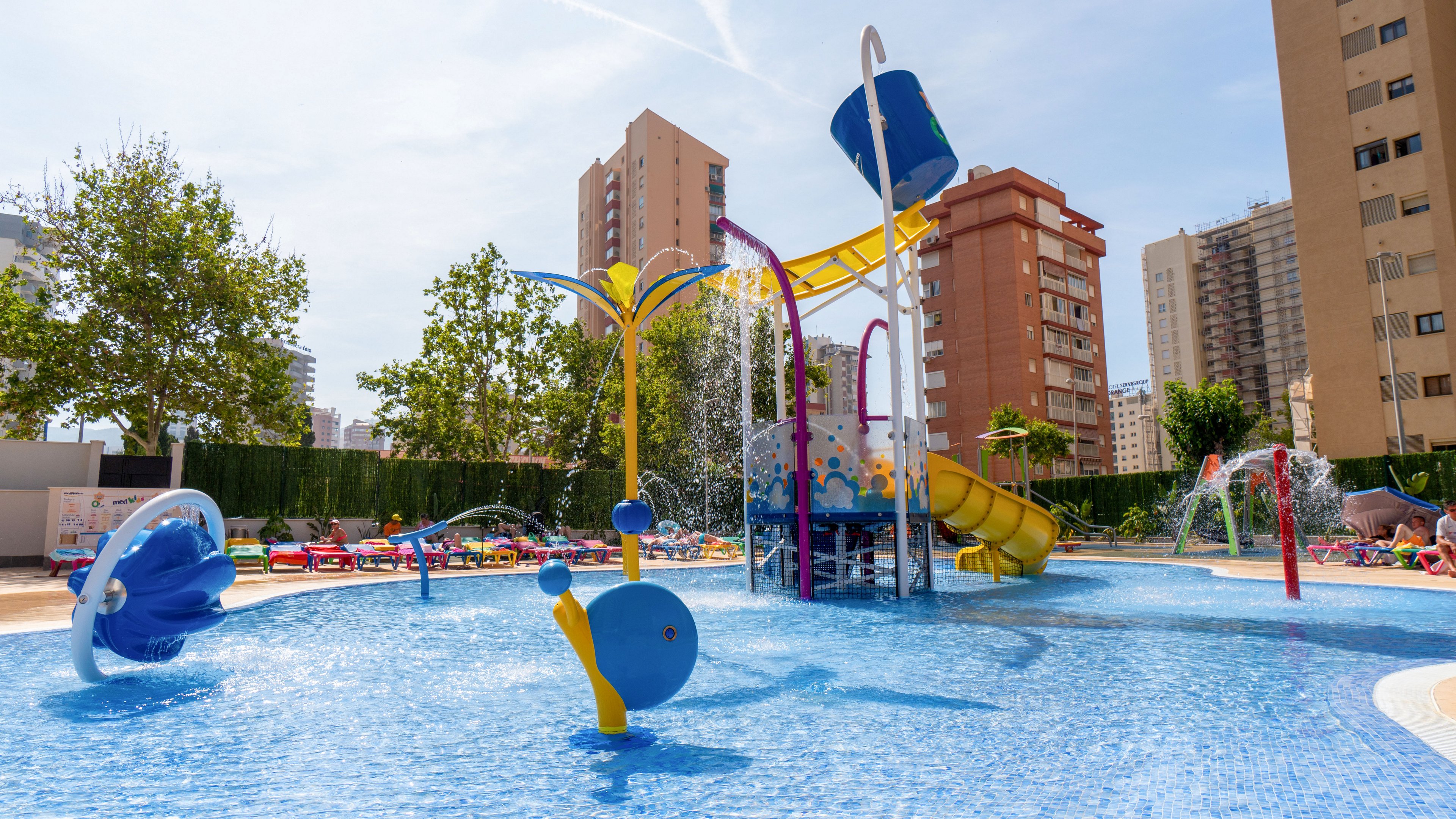 A vibrant children’s water playground features slides, splash buckets, and playful fountains in a shallow blue pool. Colorful structures and animal-shaped sprayers create an inviting family-friendly space. The scene is set outdoors on a sunny day, surrounded by lounge chairs, trees, and tall apartment buildings. Water sprays and splashes add motion and fun to the bright urban resort environment.