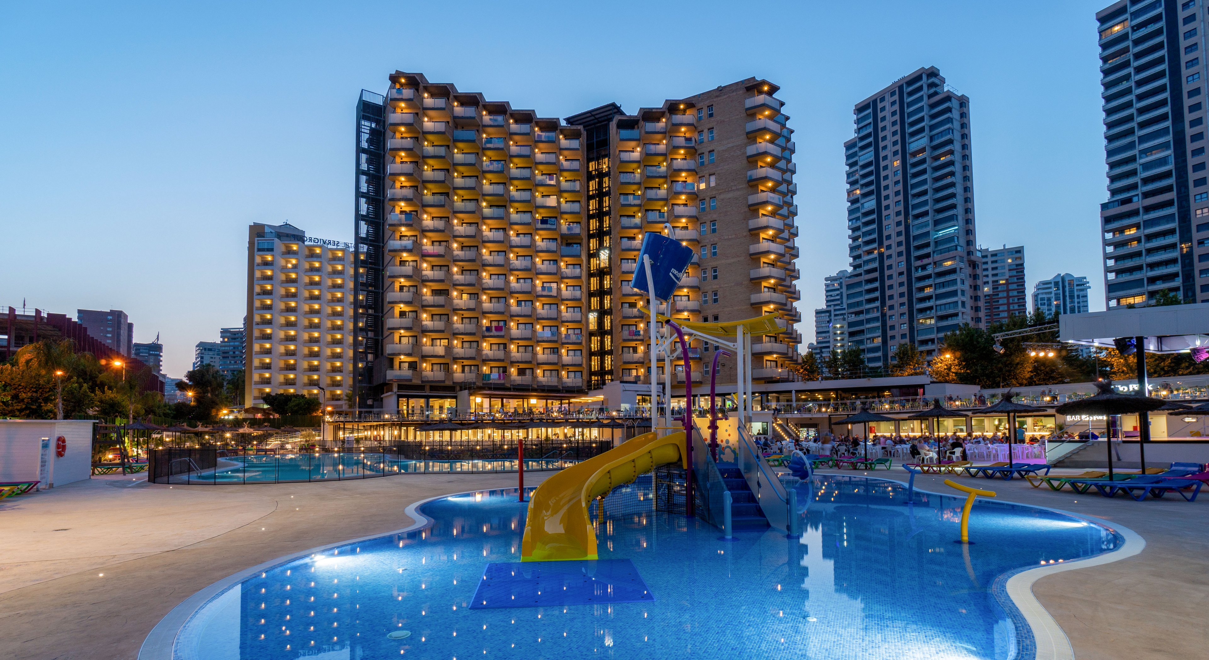 A large modern hotel building is brightly lit at dusk, with numerous windows glowing against the evening sky. In the foreground, a shallow swimming pool features colorful water slides and play structures designed for children. Surrounding the pool are lounge areas and urban high-rise buildings, creating a lively city resort atmosphere. The calm blue water reflects the lights from the hotel and nearby structures, enhancing the vibrant yet relaxed mood.