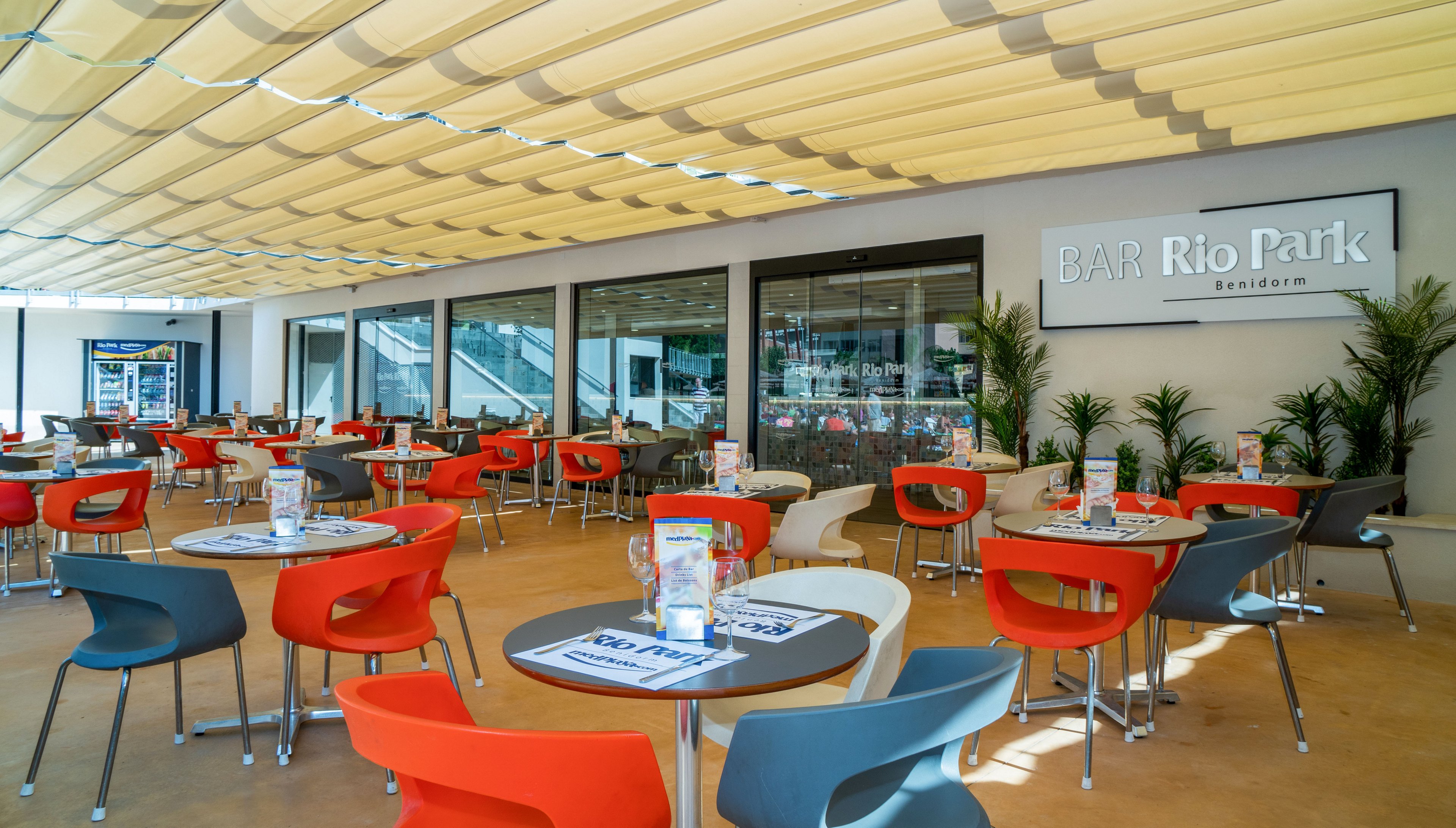Bright outdoor terrace of Bar Rio Park with colorful chairs and round tables neatly arranged. The setting appears to be part of a hotel or resort, with large glass doors and potted plants along the wall. Sunshades cover the area, creating a comfortable atmosphere for guests to sit and relax. Table menus and drink glasses add detail to the casual dining environment.