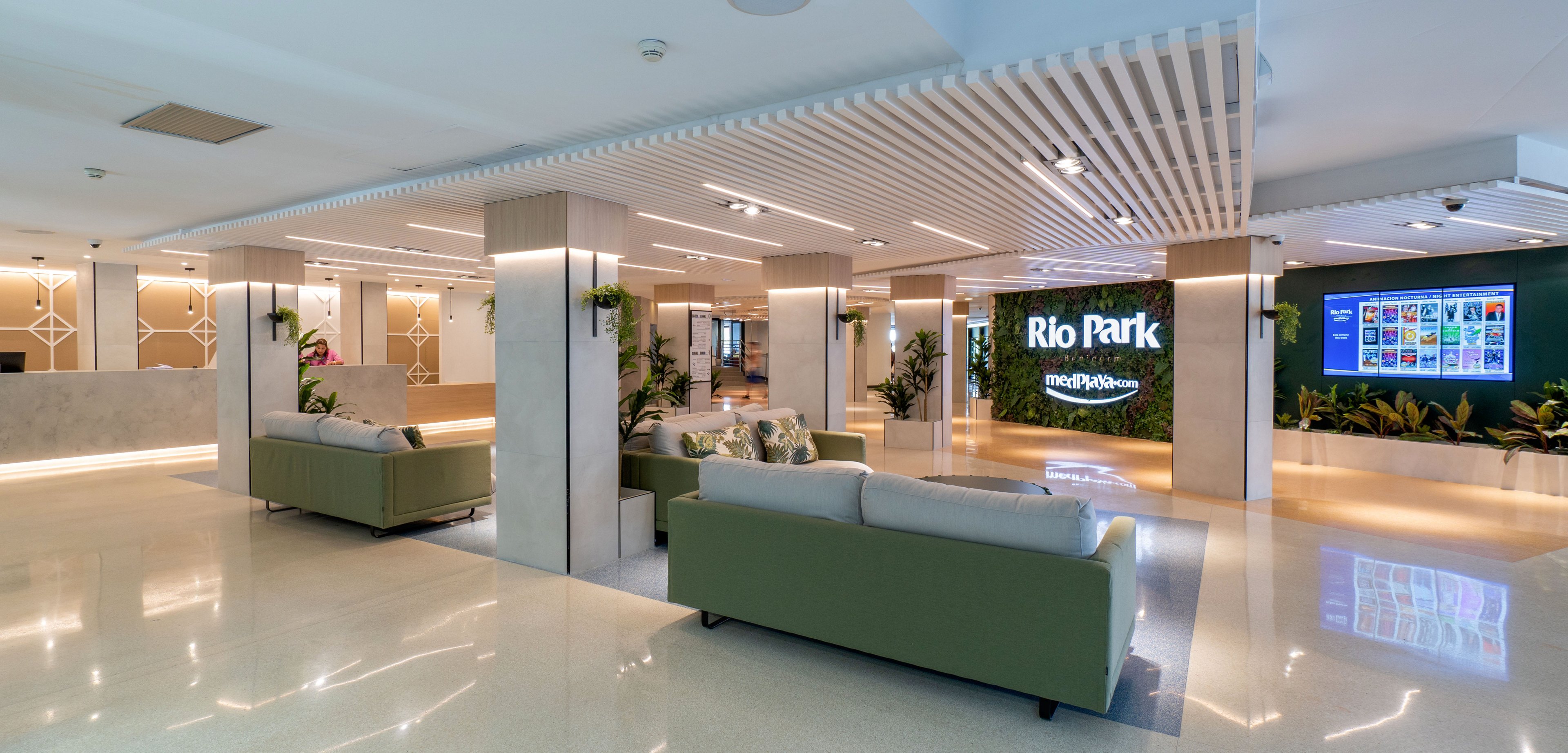 Spacious contemporary lobby area featuring multiple seating zones with green sofas and polished flooring. The setting includes decorative wall panels, indoor plants, and warm ambient lighting that creates an inviting atmosphere. A large digital display clearly shows the text "Rio Park" alongside additional signage and a screen with colorful content. The overall style is clean, modern, and well-lit, suitable for hospitality or corporate environments.