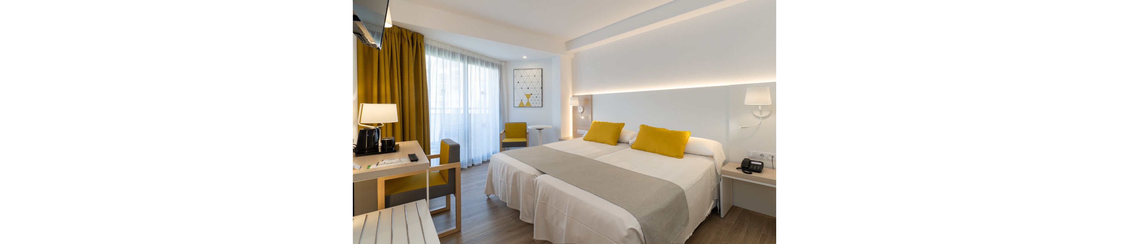 Bright contemporary hotel room featuring two neatly made twin beds with yellow accent pillows and a light gray runner. The space includes a desk area with a coffee station, chair, and wall-mounted television. Large floor-to-ceiling windows with sheer curtains and mustard yellow drapes allow natural light to fill the room. Minimalist decor, soft lighting, and neutral tones create a clean, welcoming atmosphere.
