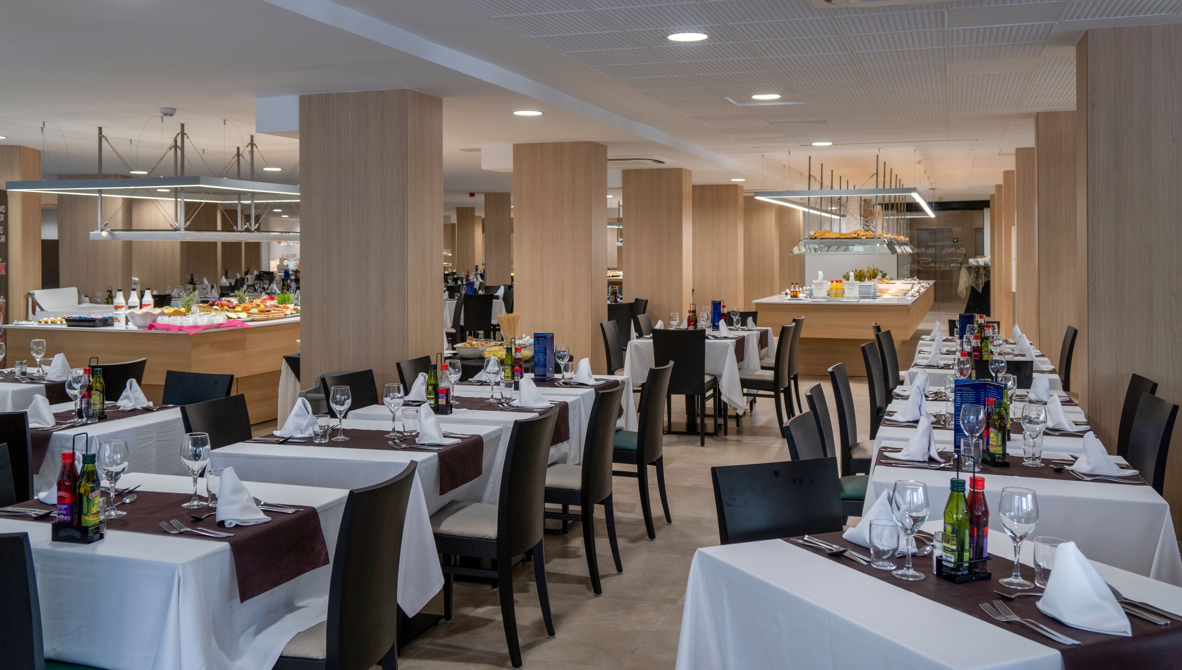 Spacious contemporary restaurant dining room with neatly arranged tables and chairs. White tablecloths, folded napkins, glassware, and condiments create an elegant yet casual atmosphere. In the background, a brightly lit buffet area displays various dishes under modern fixtures. Light wood columns and neutral tones give the space a clean, modern design. No people are present, emphasizing the prepared setting ready for guests.